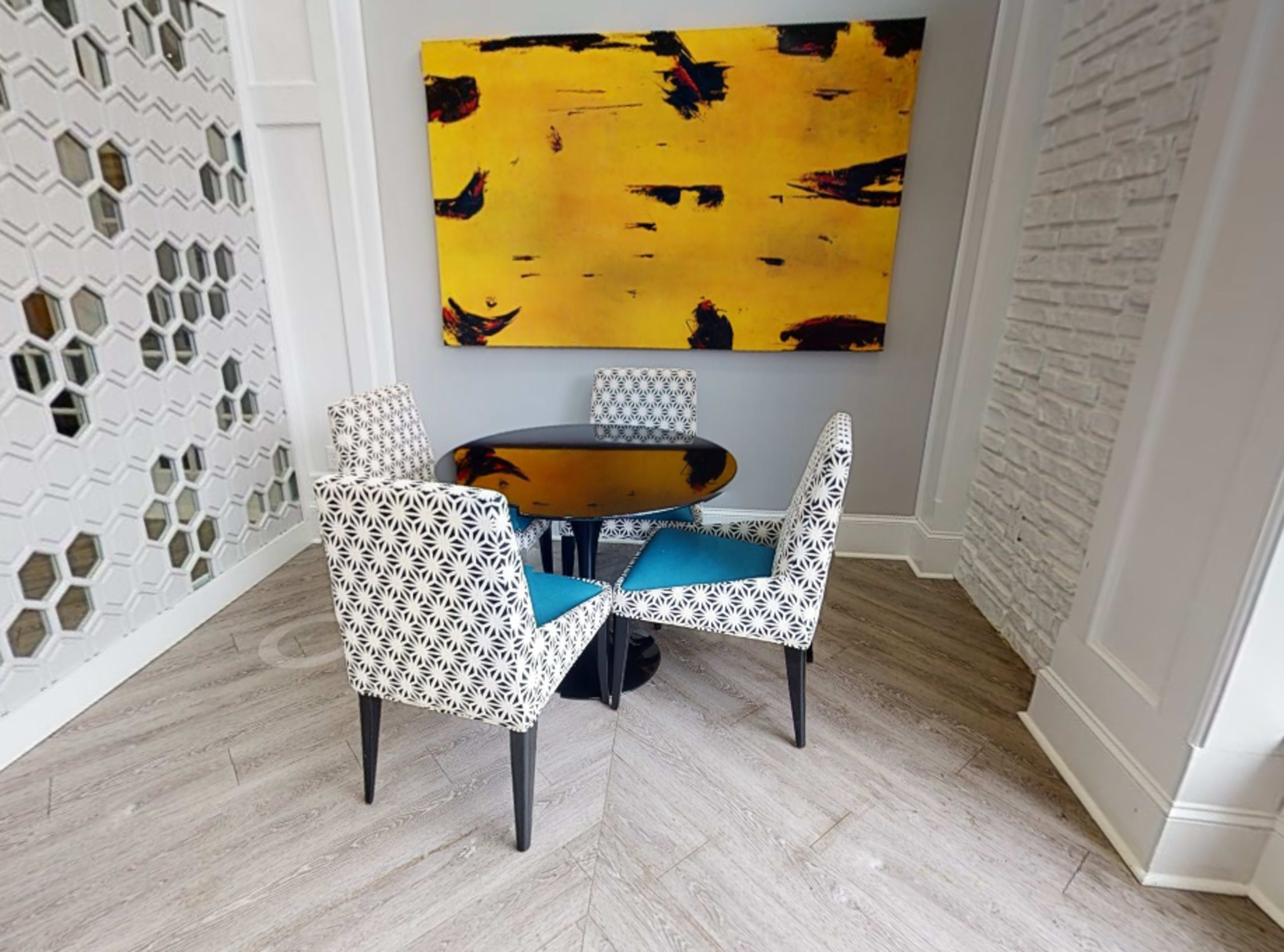 A small dining table with four patterned chairs is positioned near a large abstract painting on the wall in a brightly lit corner.