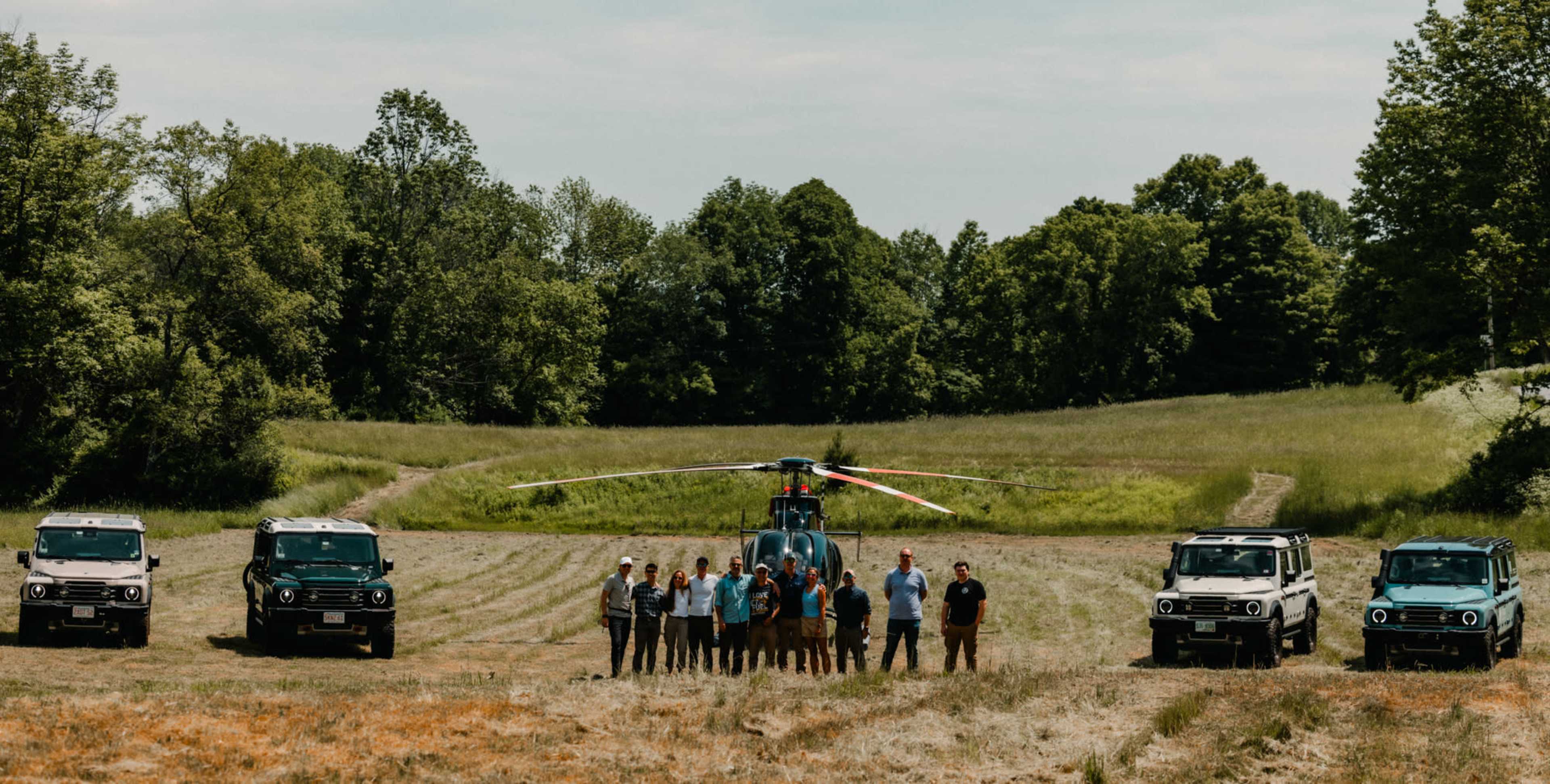 A group of people stands in a field surrounded by four vehicles and a helicopter with trees in the background.