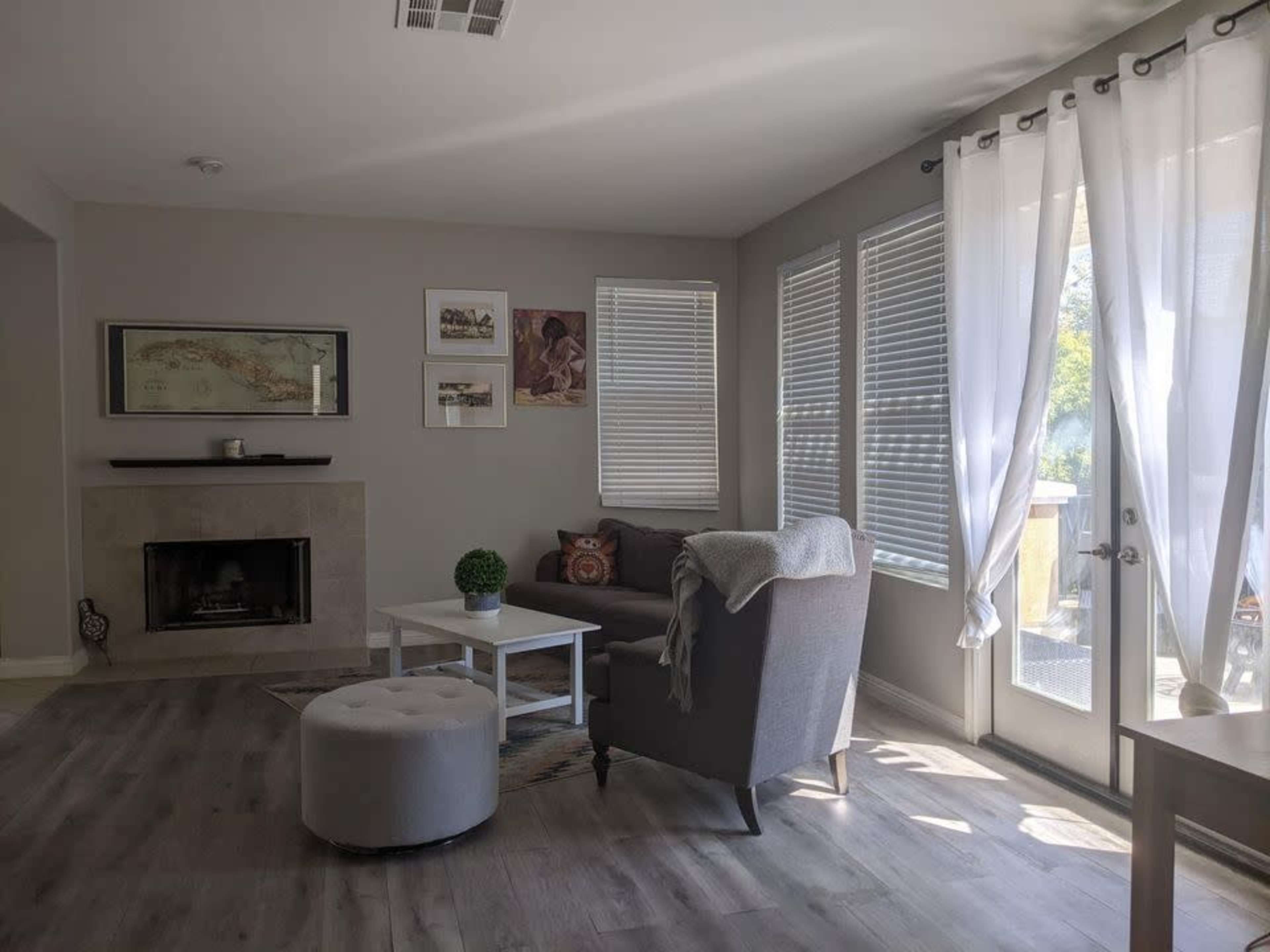 A light-filled living room features a gray armchair, a small round ottoman, a white coffee table, and a fireplace, with large windows and sliding glass doors dressed in sheer curtains.