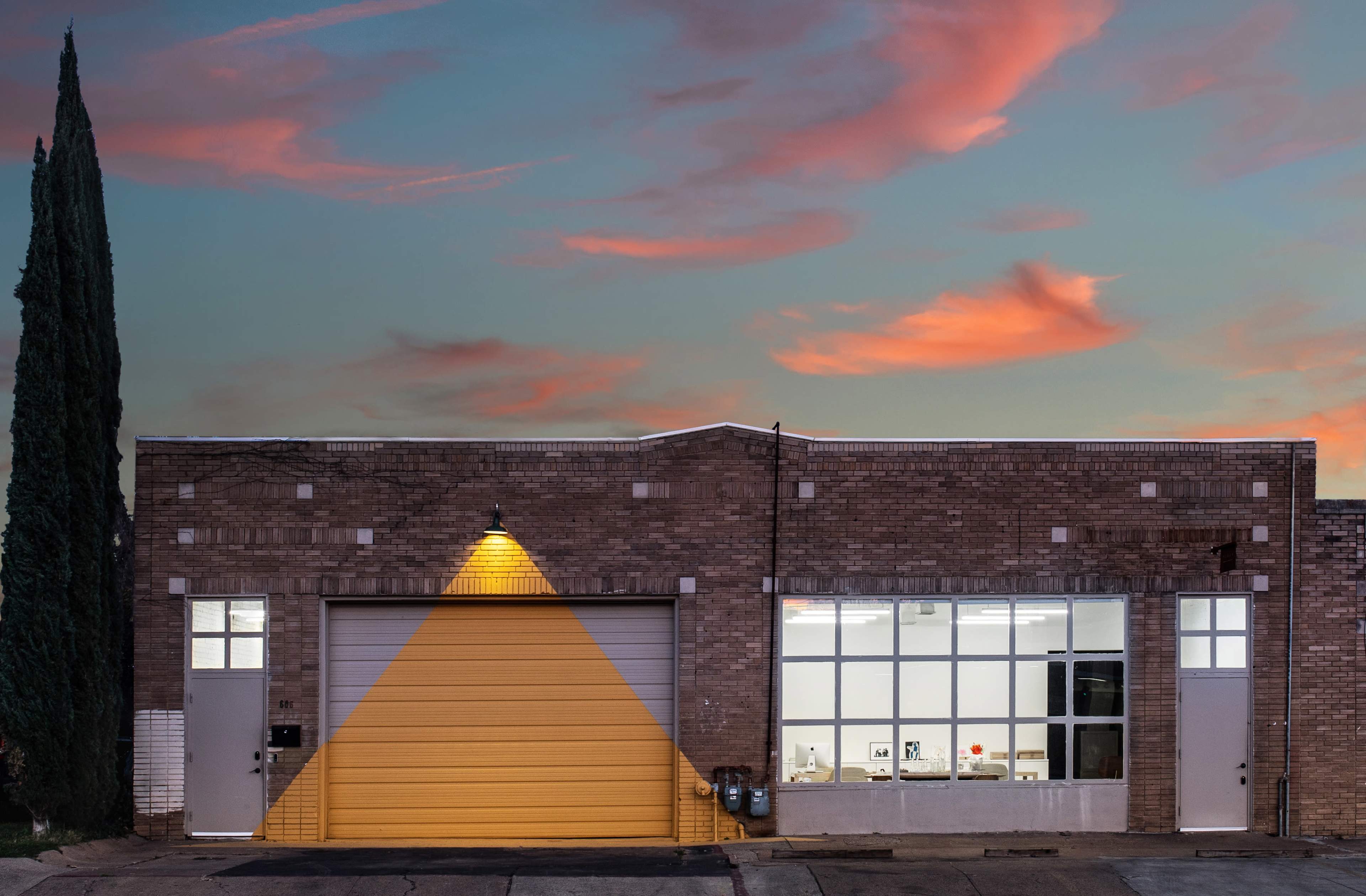 The image shows a brick building with a large orange garage door, a window displaying a lit interior, and a tall tree beside it against a colorful sunset sky.