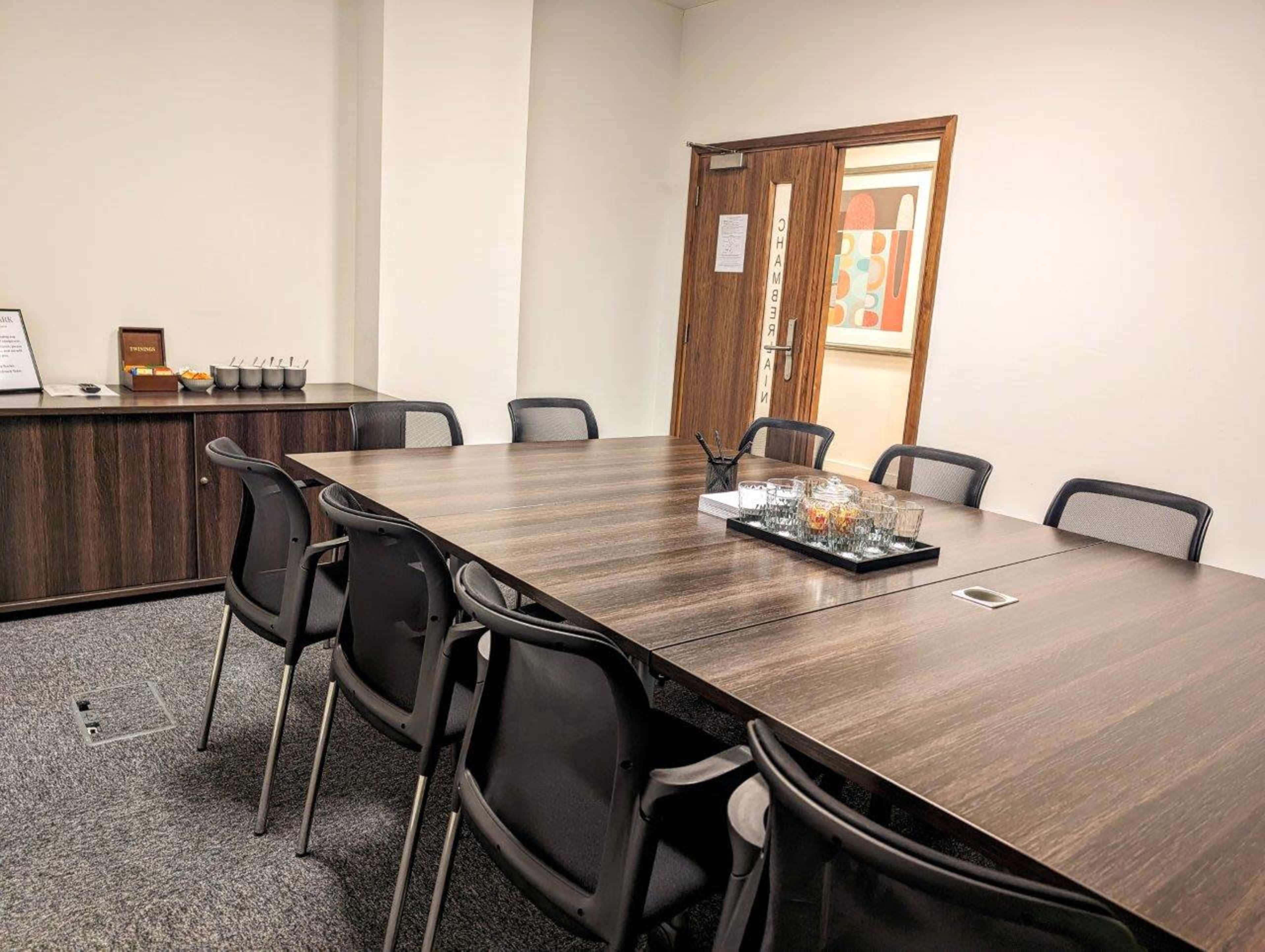 A conference room features a large brown table surrounded by black chairs, with a tray of glasses and a decorative bowl on the table, and a wooden cabinet against the wall.