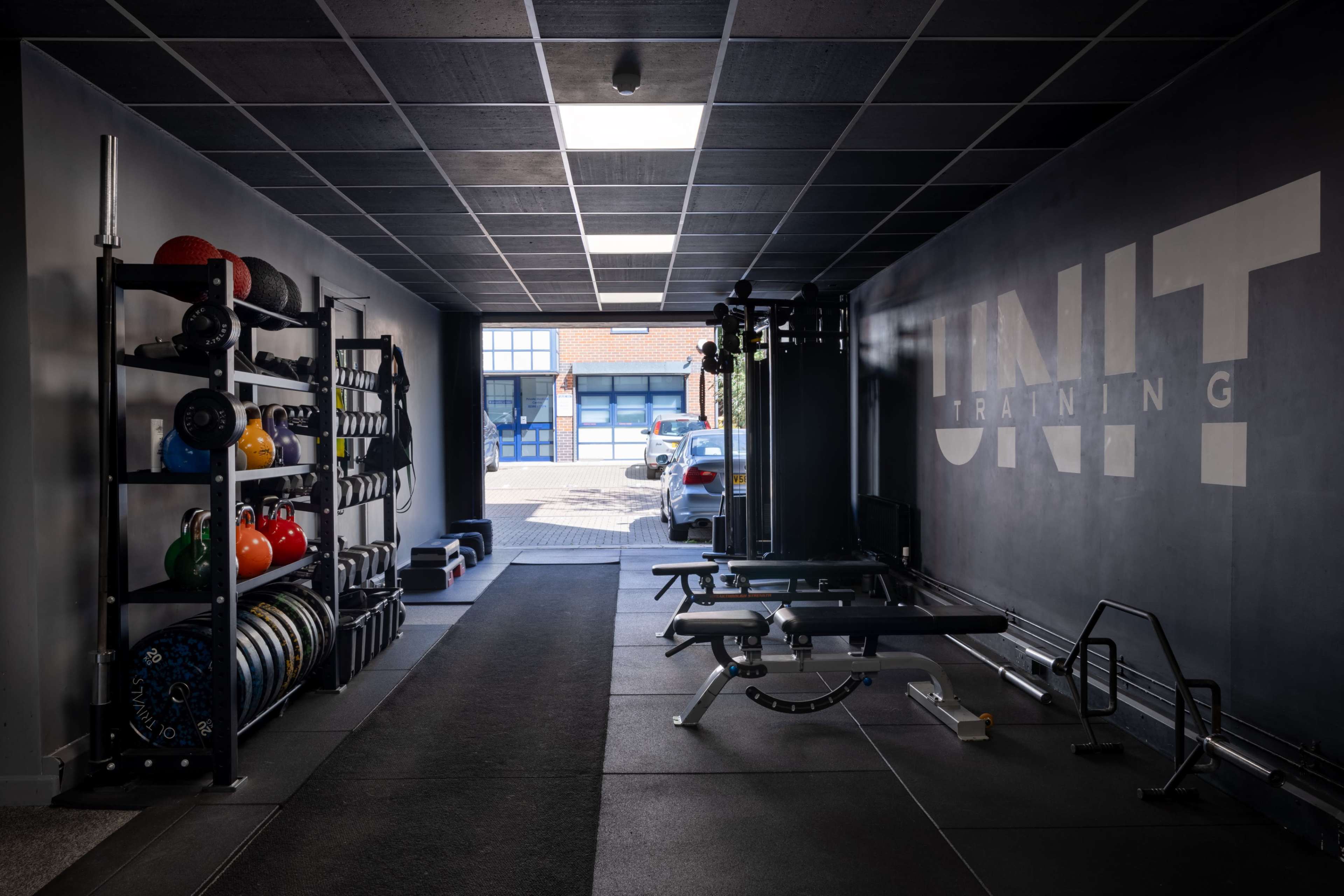 The image shows a modern gym interior featuring a variety of fitness equipment, including weights on a rack and a workout bench, with large windows visible at the far end.