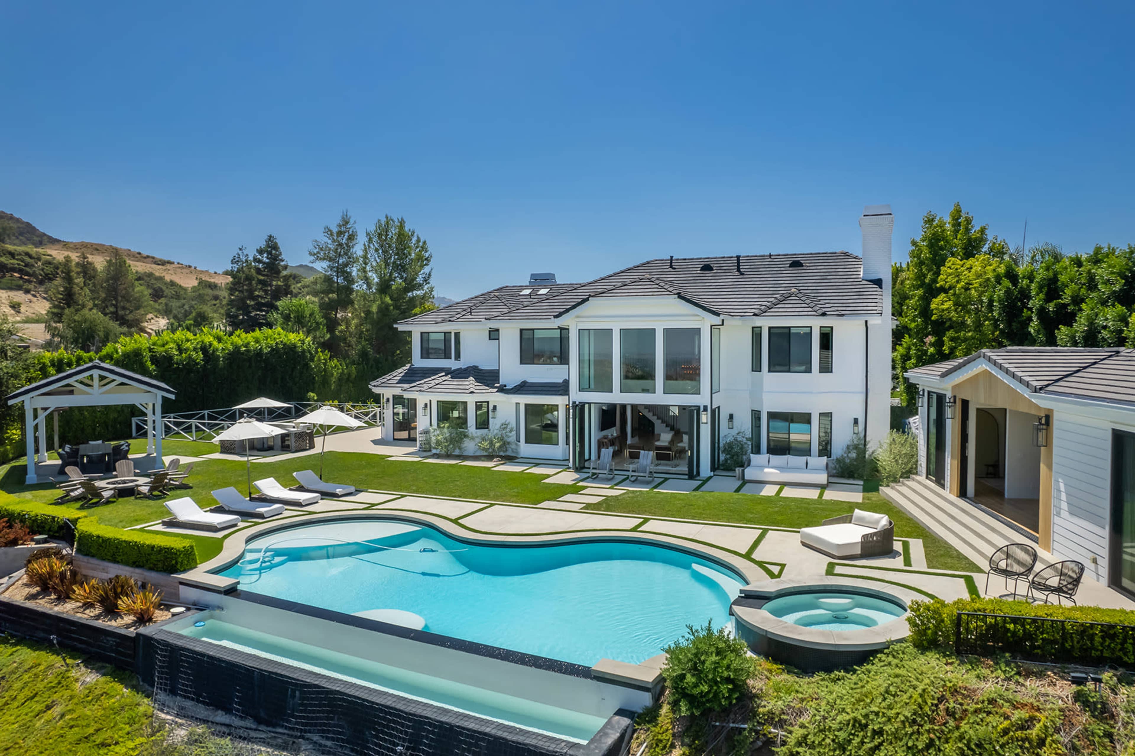 A large white two-story house with a swimming pool and landscaped yard, surrounded by trees and outdoor seating areas.