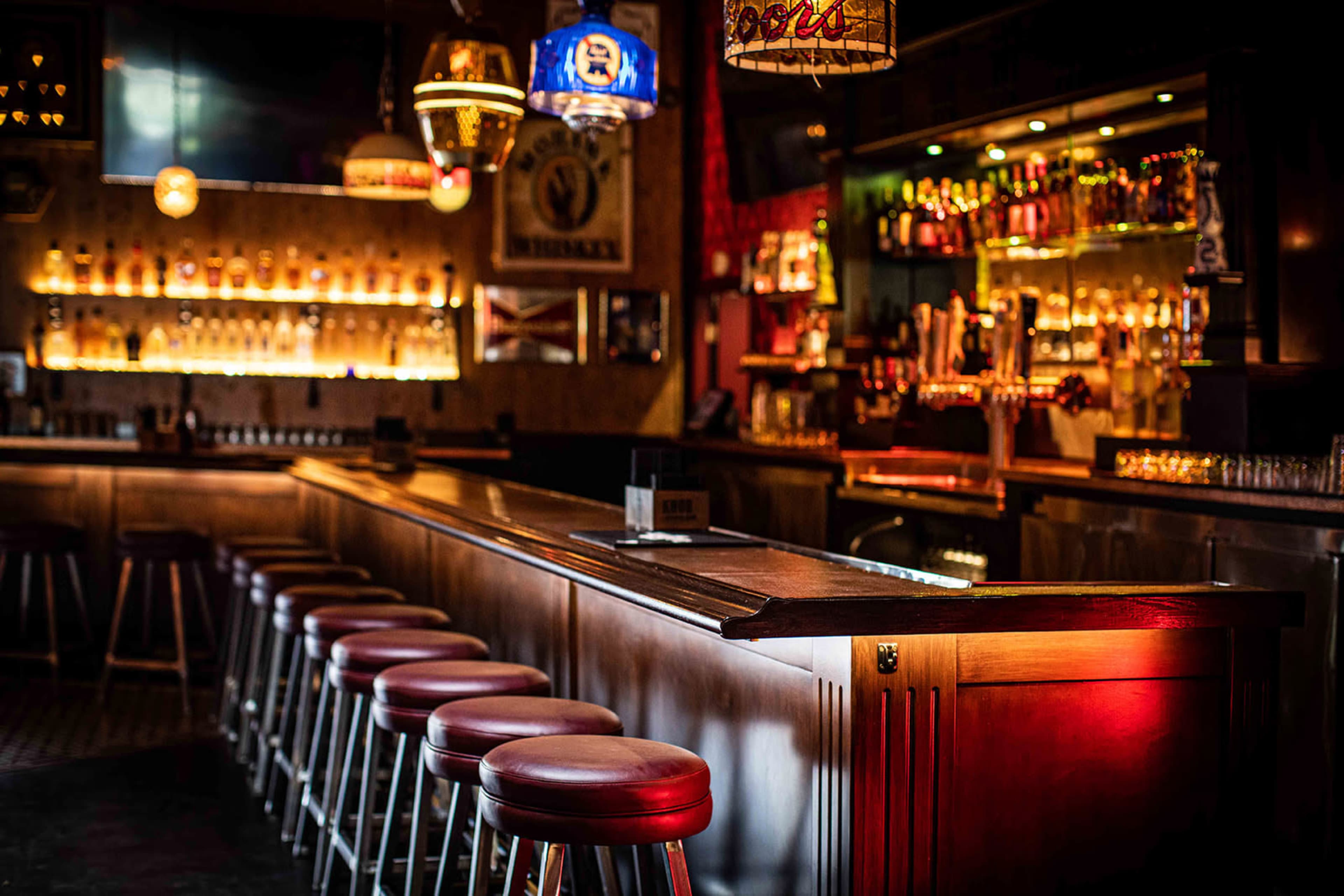 The image shows a dimly lit bar with wooden stools lining a polished countertop and shelves stocked with various bottles.