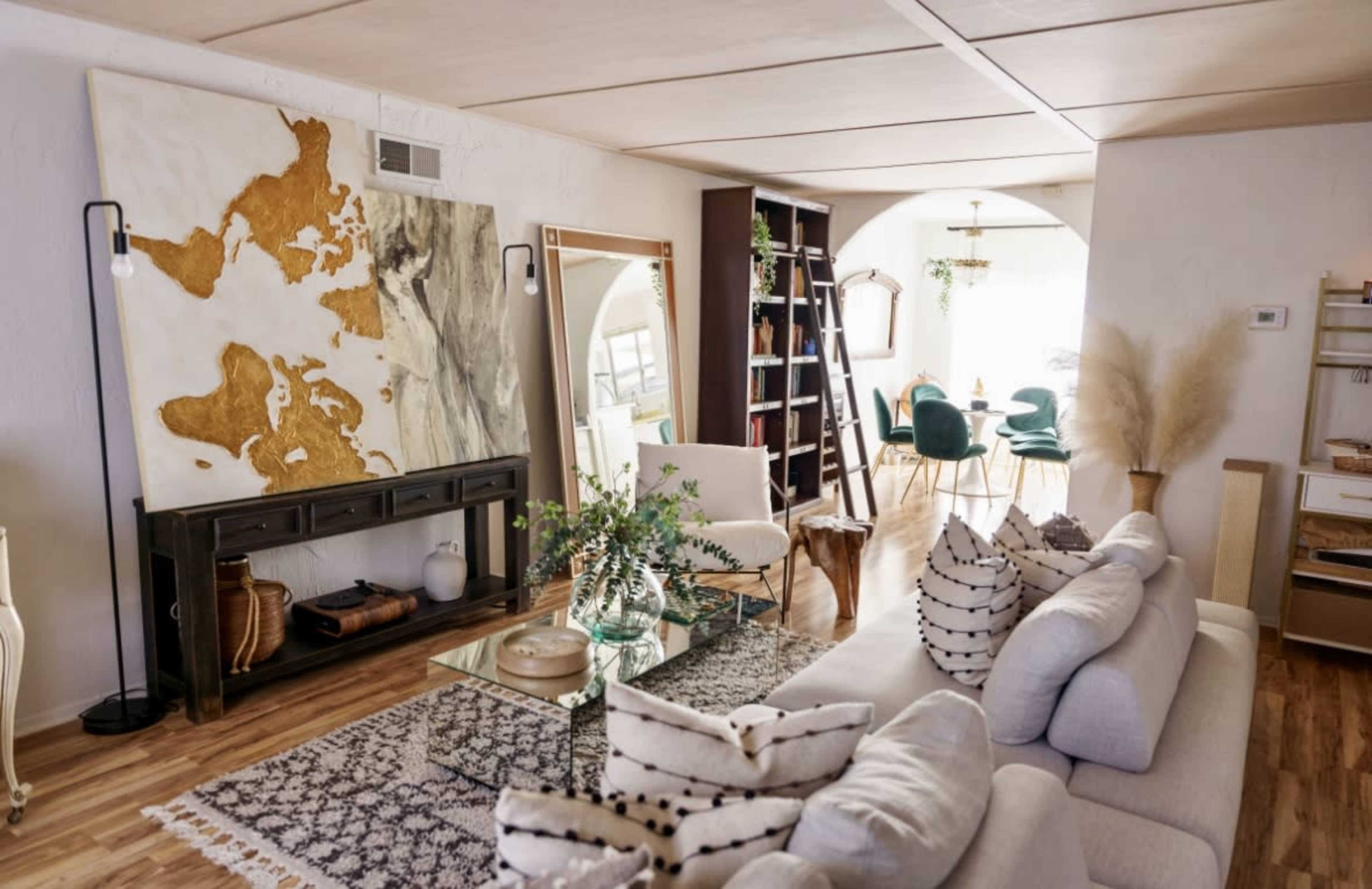 A modern living room featuring a large abstract map painting, a sectional sofa, a glass coffee table, and a dining area in the background with teal chairs.