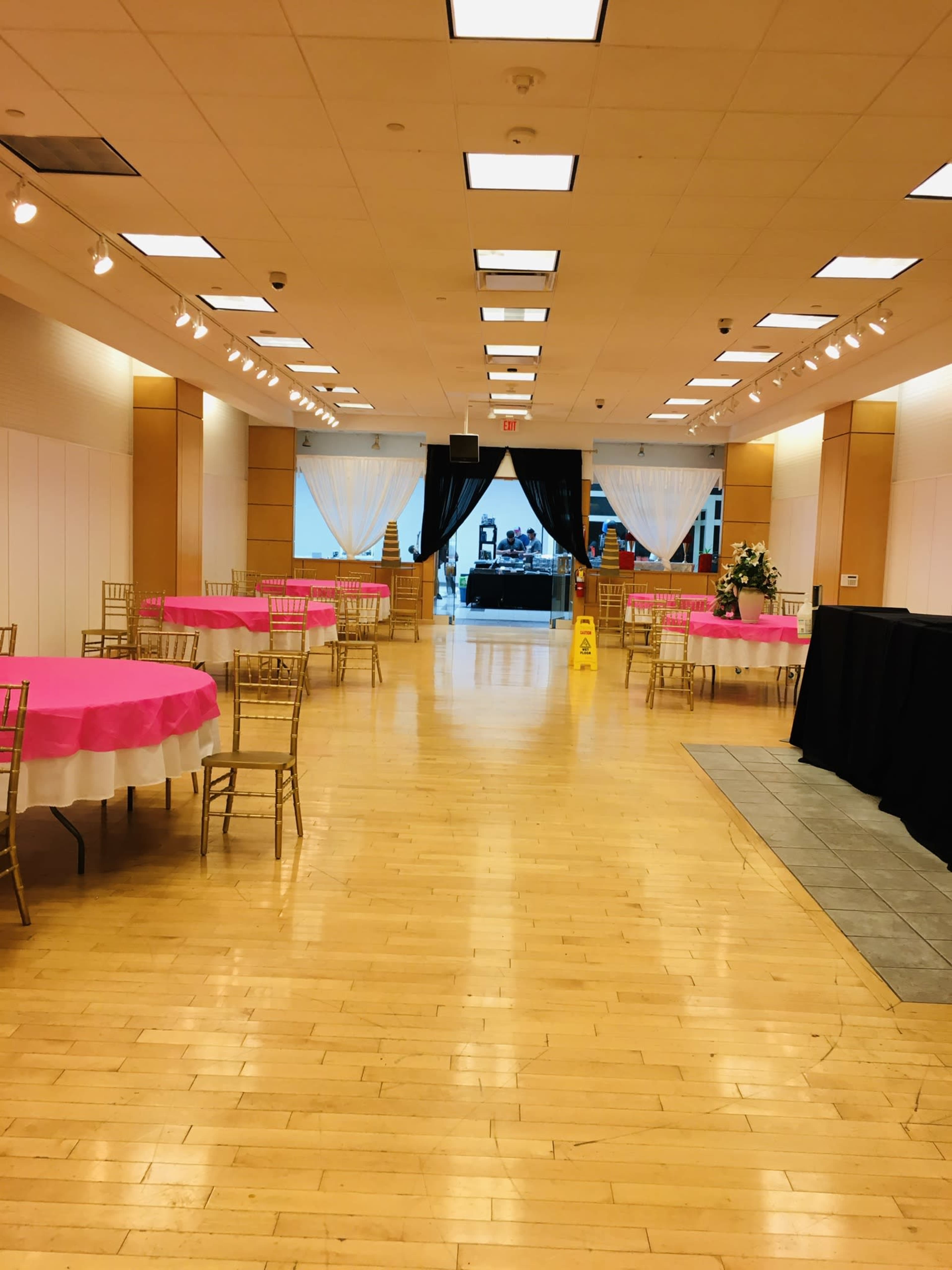 A banquet hall with round tables covered in pink tablecloths, a polished wooden floor, and a stage area at the far end.