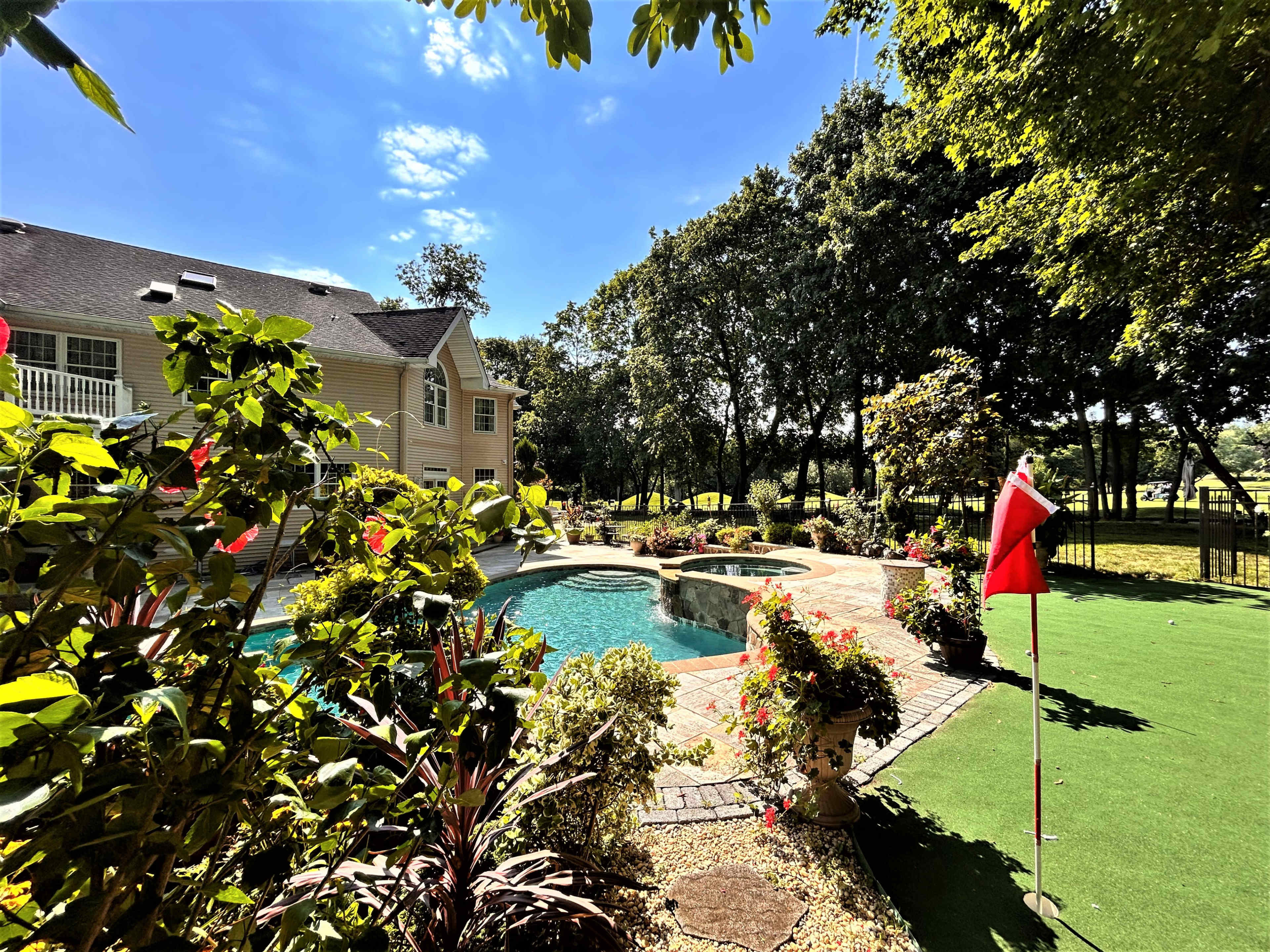 The scene shows a landscaped backyard with a swimming pool, spa, and a golf putting green, surrounded by trees and colorful flowers.