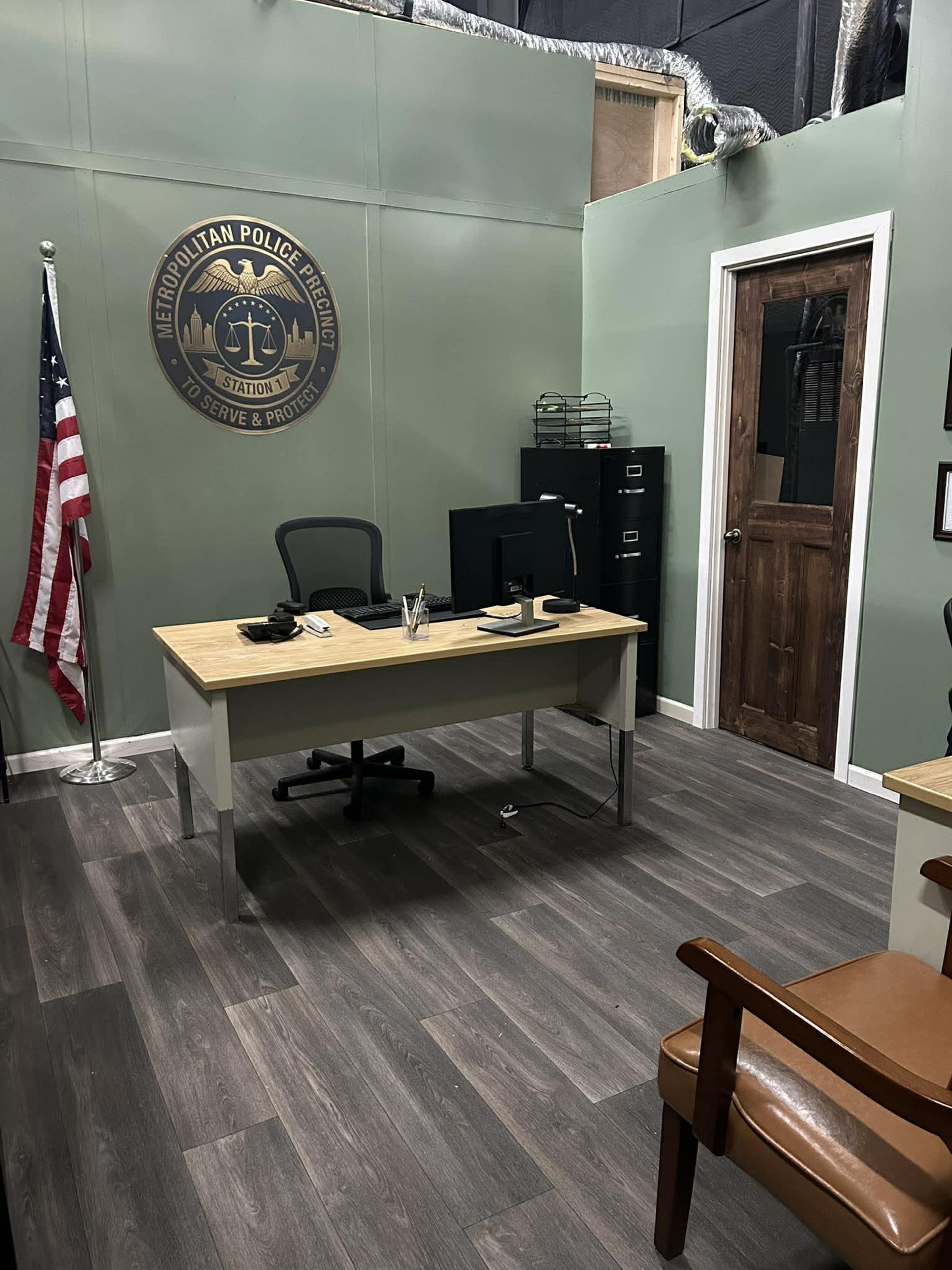 The image shows a police office featuring a desk with a computer, a chair, an American flag, a door, and a wall-mounted emblem of a police department.