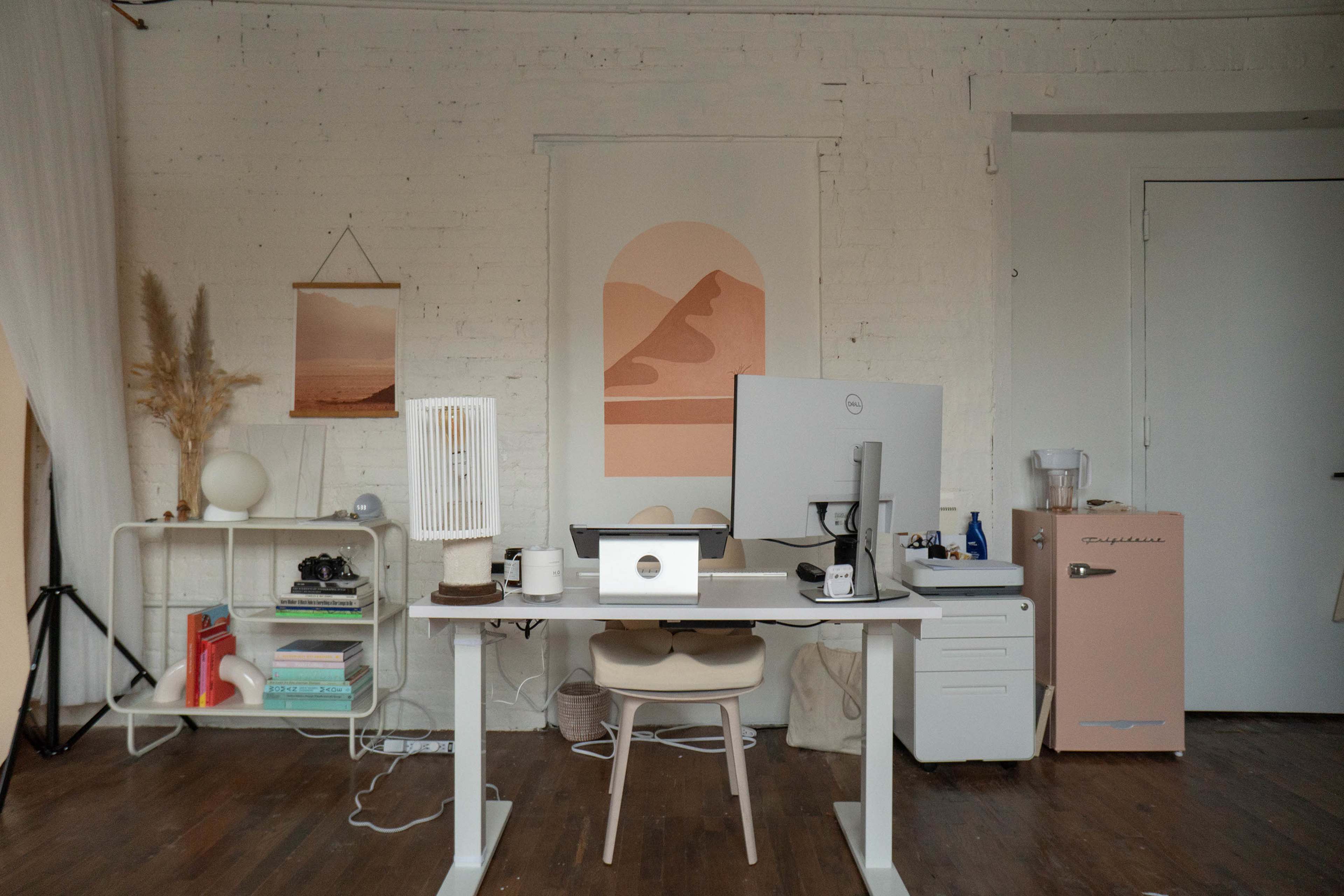 A minimalist office setup featuring a desk with a computer monitor, a chair, and decorative elements against a simple white brick wall.