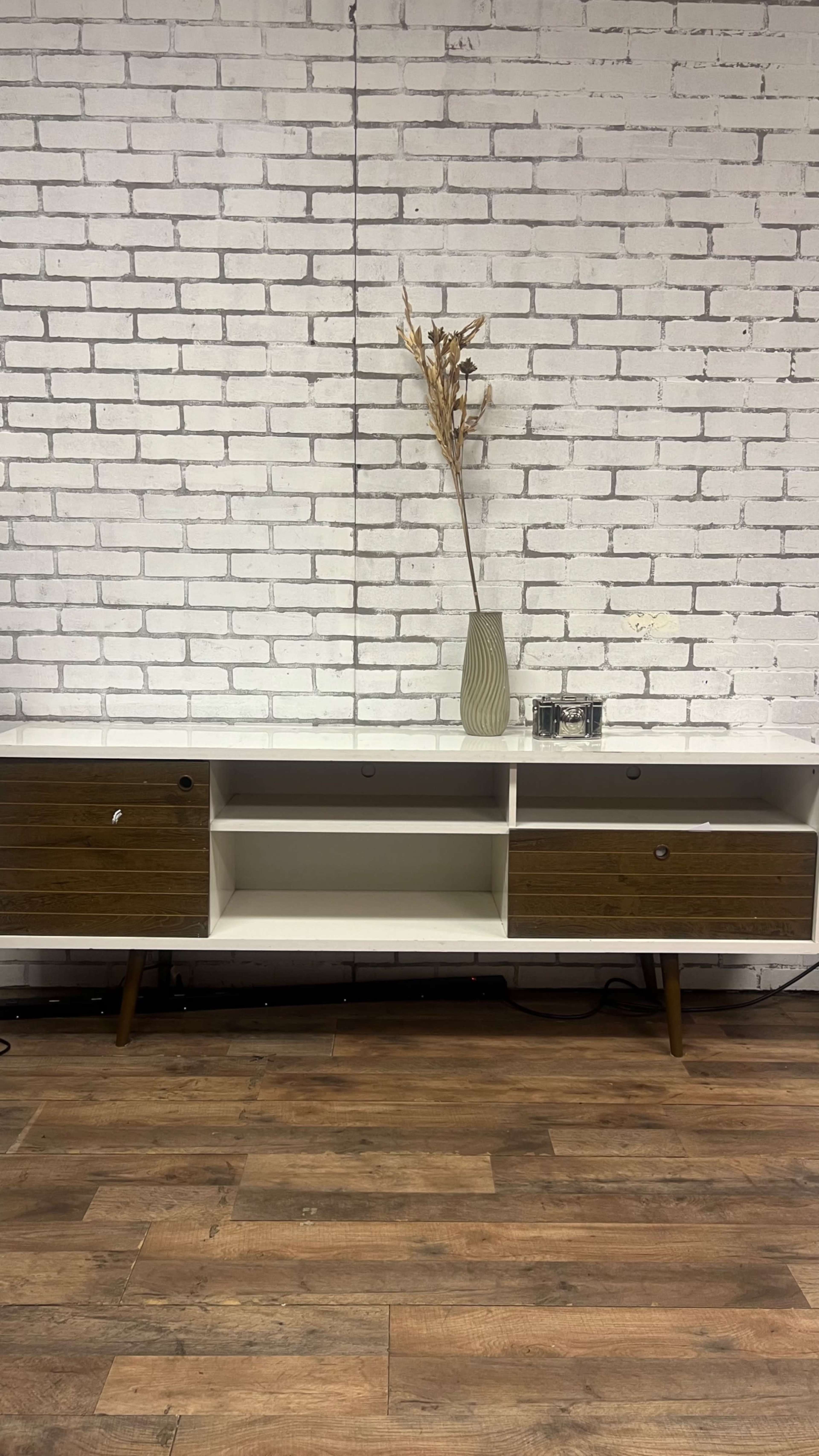 The image shows a white and brown wooden TV stand against a white brick wall, featuring two drawers, an open shelf, and a vase with dried plants on top.