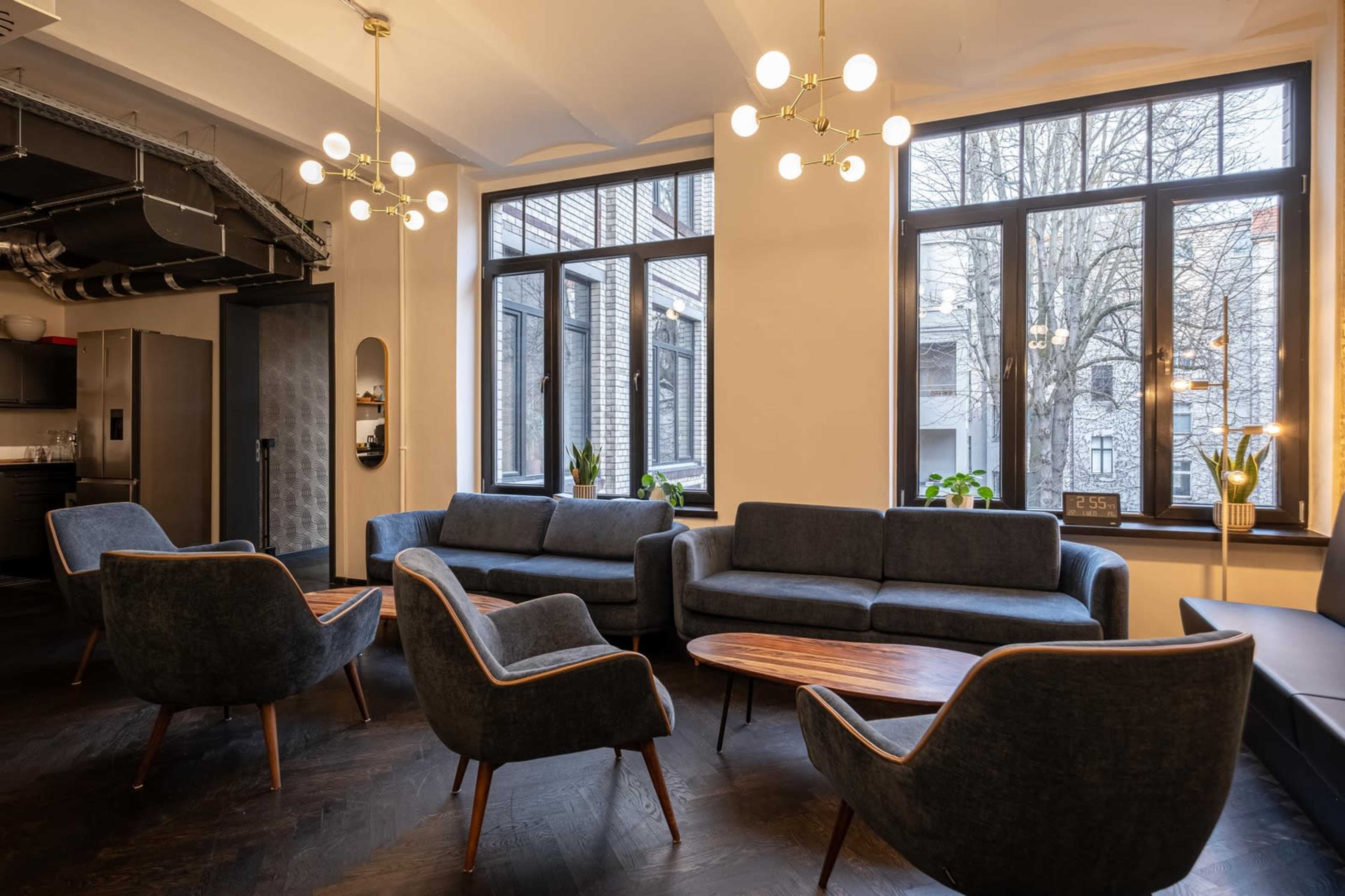 The image shows a modern lounge area with several gray sofas and armchairs arranged around a wooden coffee table, illuminated by pendant lights and natural light coming through large windows.