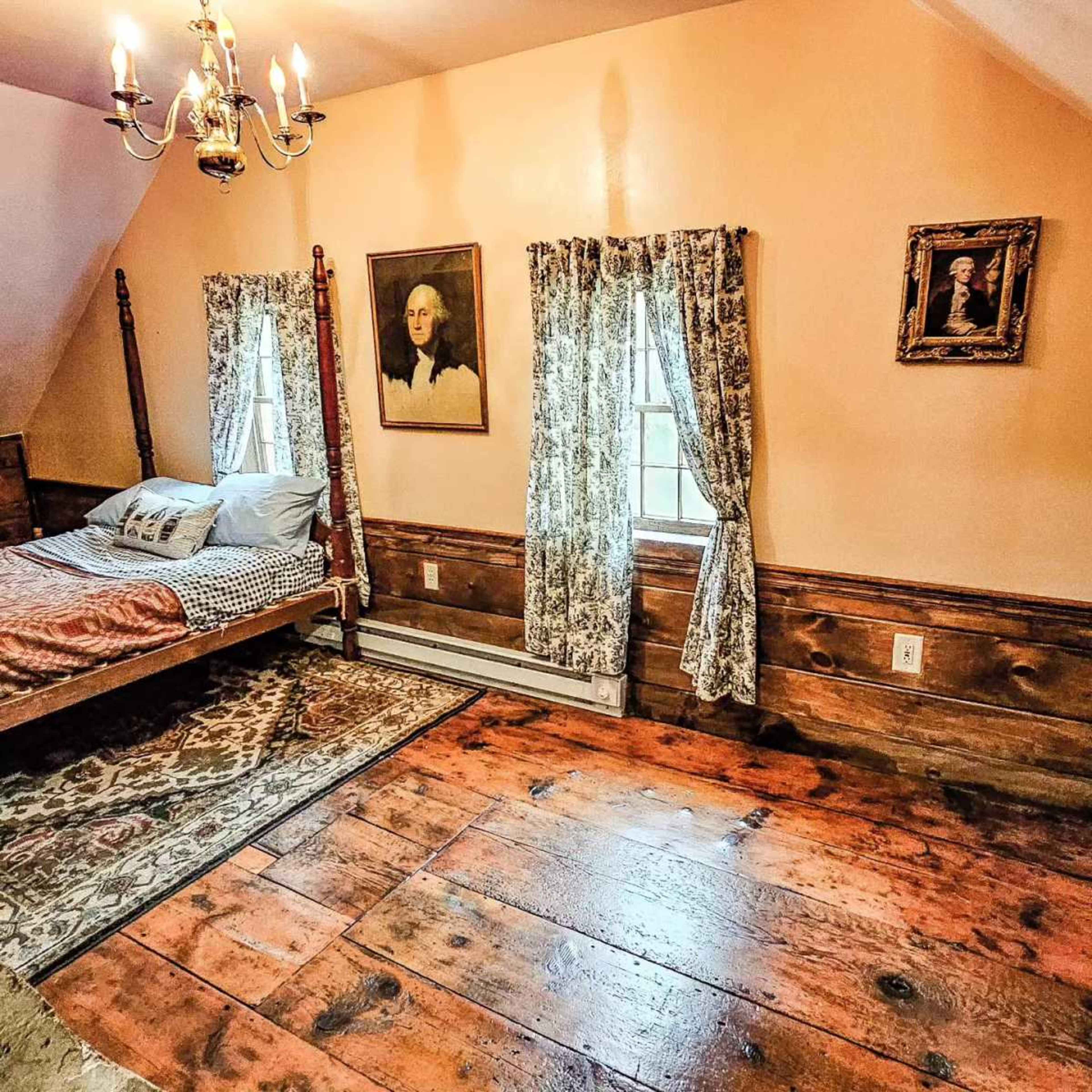 The image shows a cozy bedroom with a wooden bed, patterned curtains, and two framed portraits on the walls.