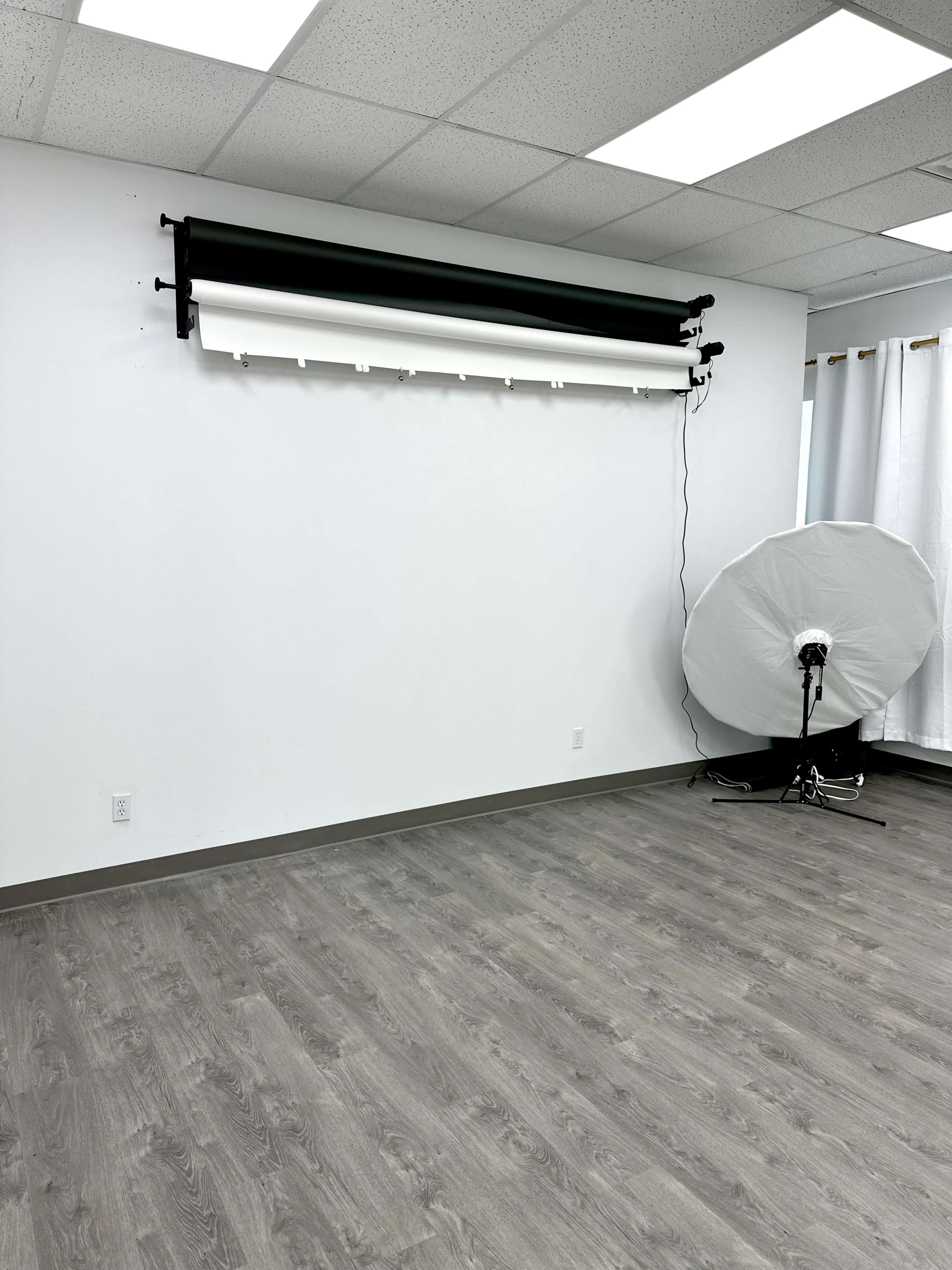 An empty room with a white wall, a roll of black and white backdrop paper mounted above, and a large round softbox light on a stand.
