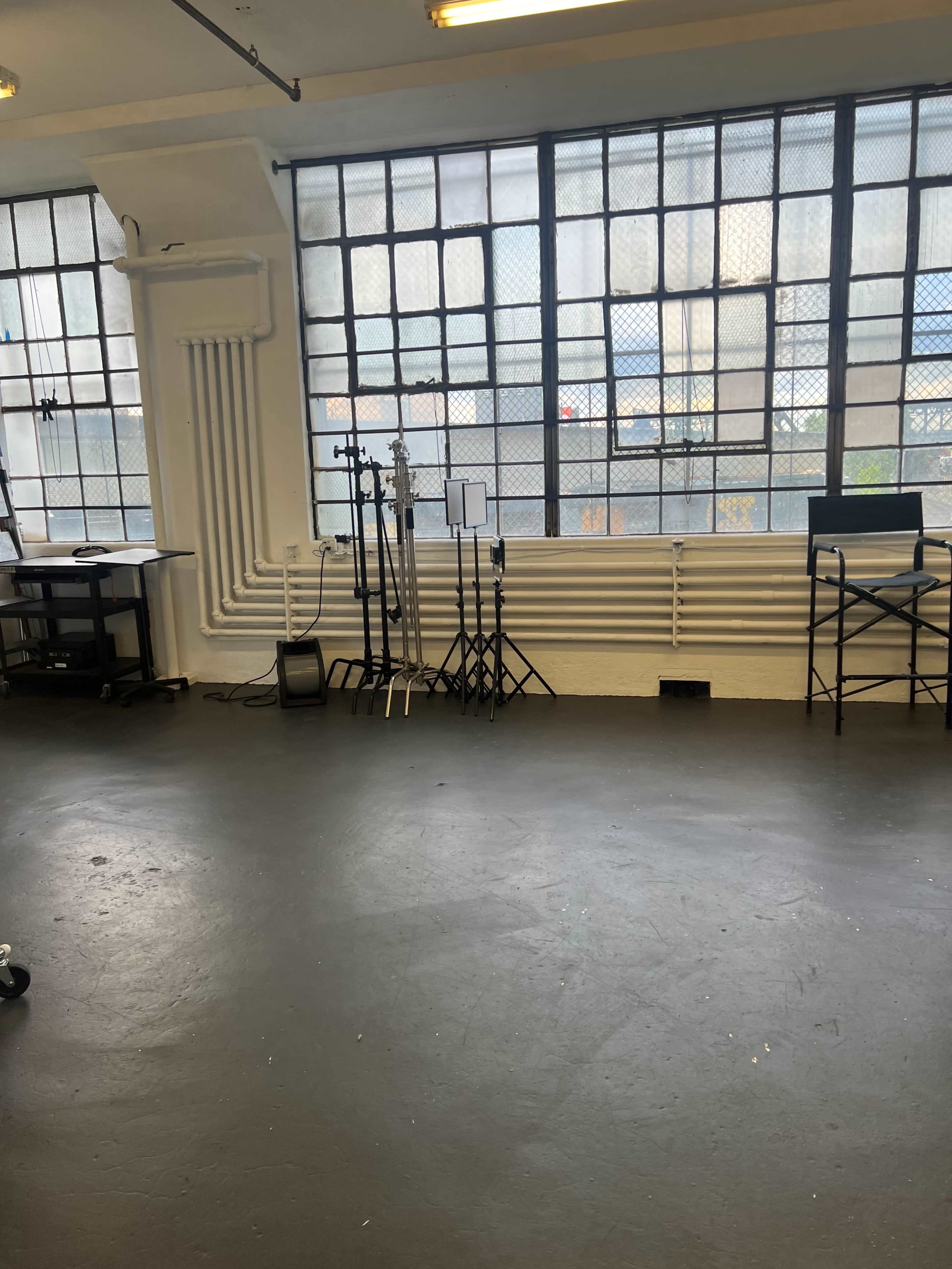 A spacious studio interior with large windows, a black floor, and various lighting stands arranged along the wall.