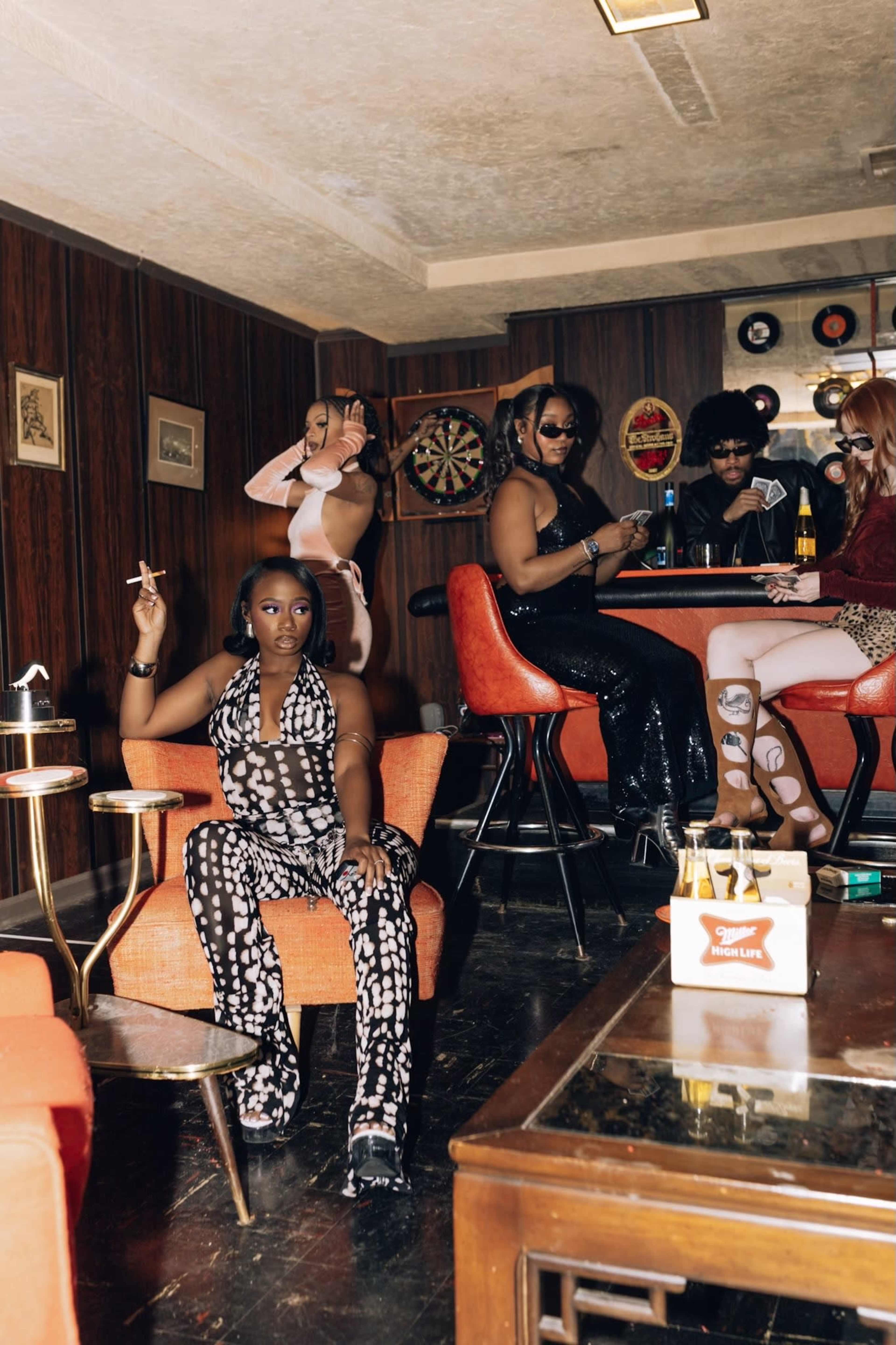 A group of five people socialize in a retro-style lounge with a bar, while one person sits casually in an orange chair smoking a cigarette.