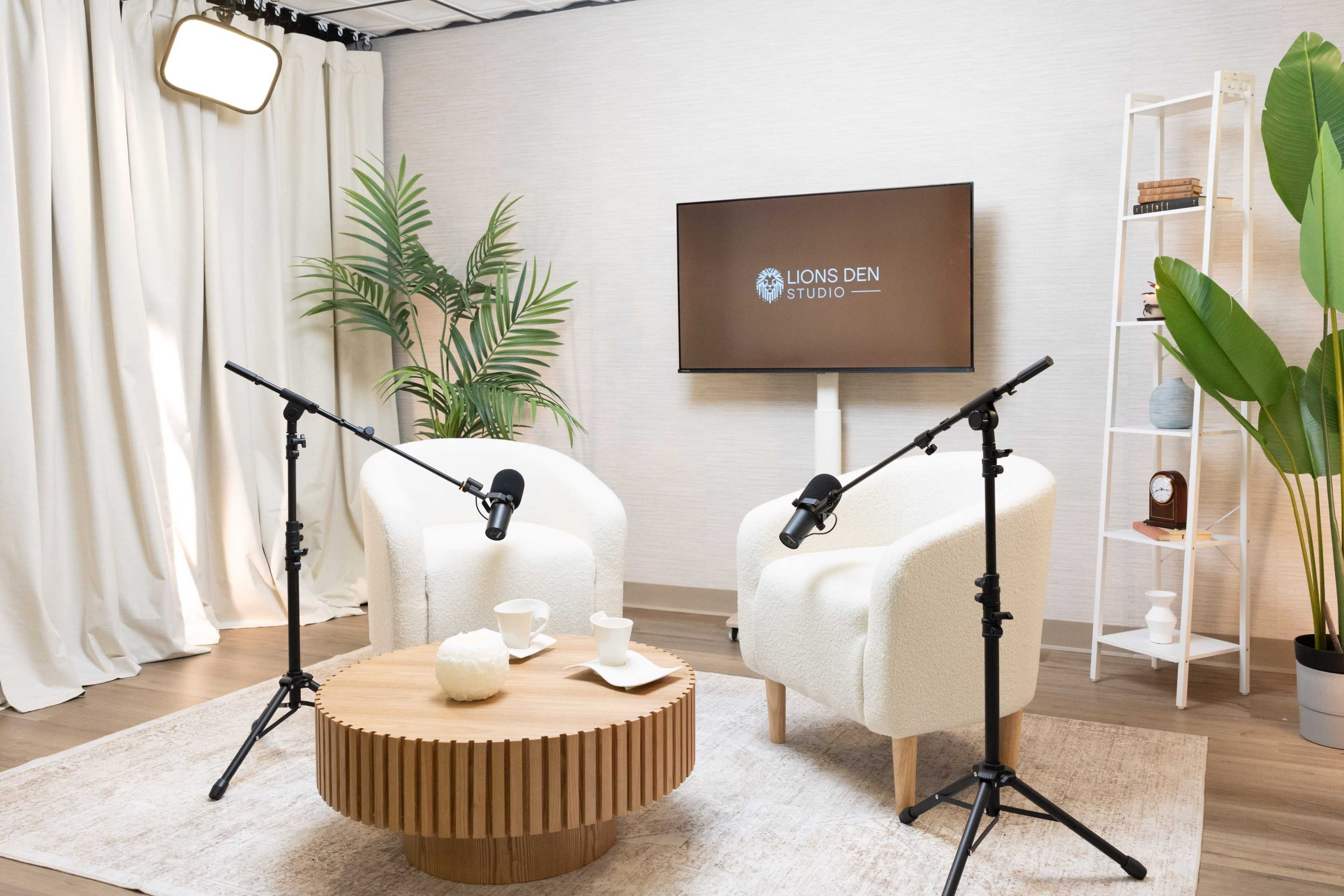 The image shows a modern studio setup with two white chairs facing a television screen, surrounded by plants and a round wooden coffee table with tea cups.