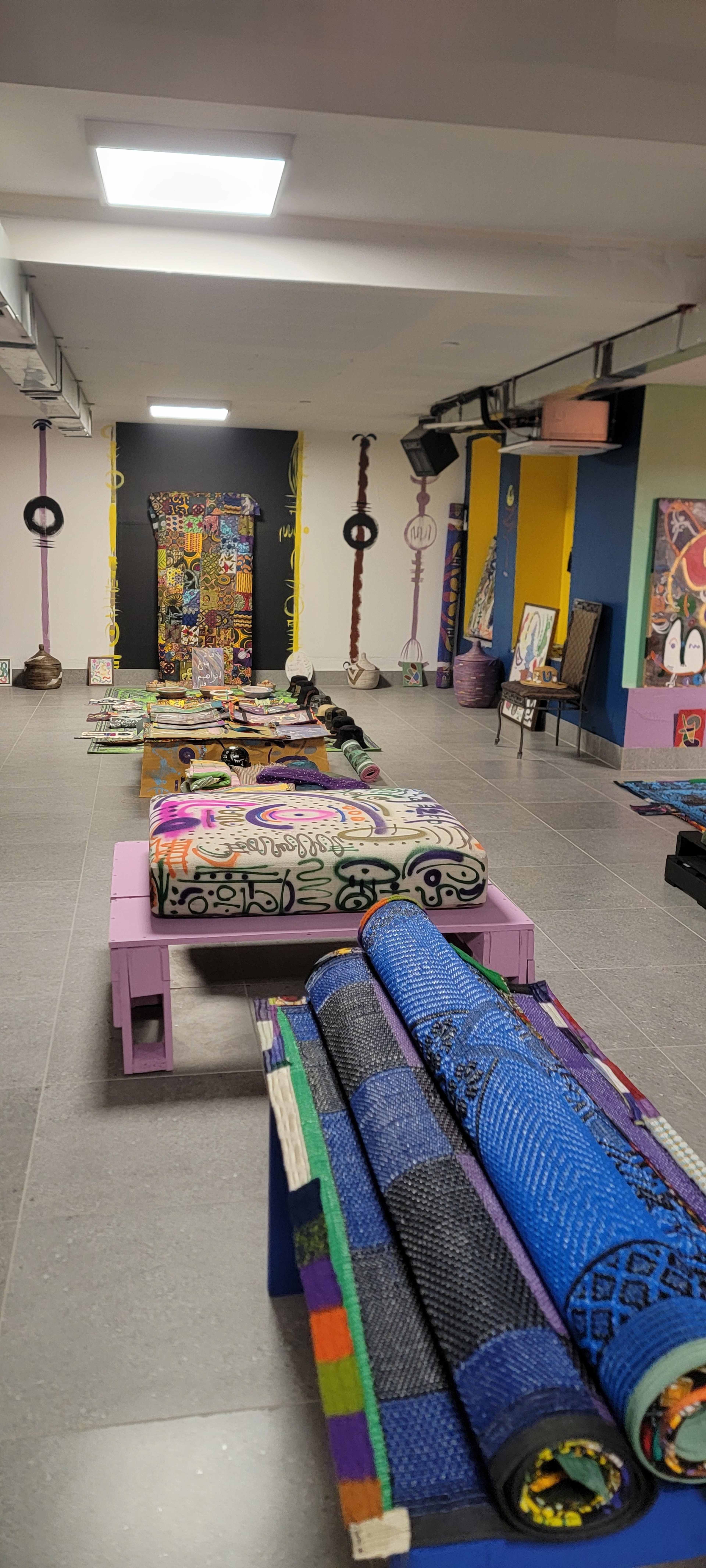 The image shows a colorful art installation featuring benches and rolled mats arranged along a spacious, well-lit room with various artworks displayed on the walls.