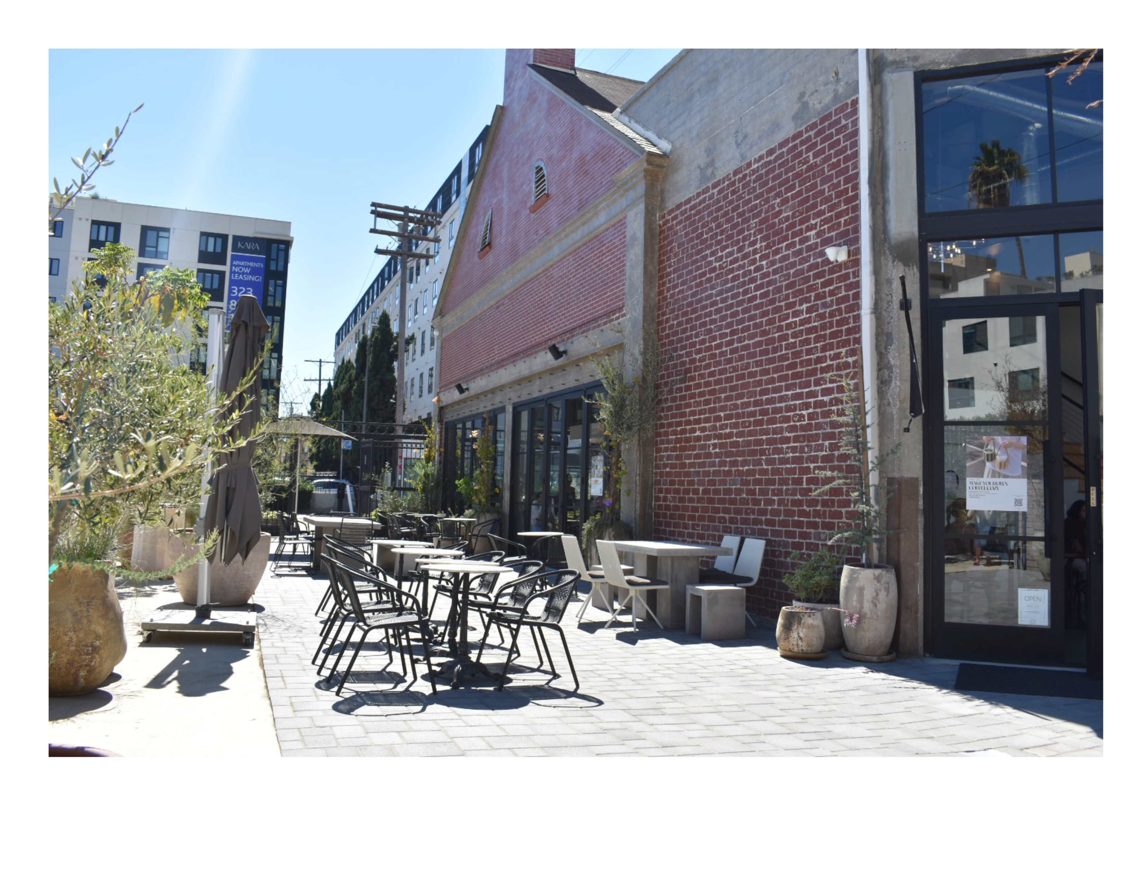 The image shows an outdoor seating area of a café or restaurant with metal tables and chairs surrounded by potted plants and a brick building.