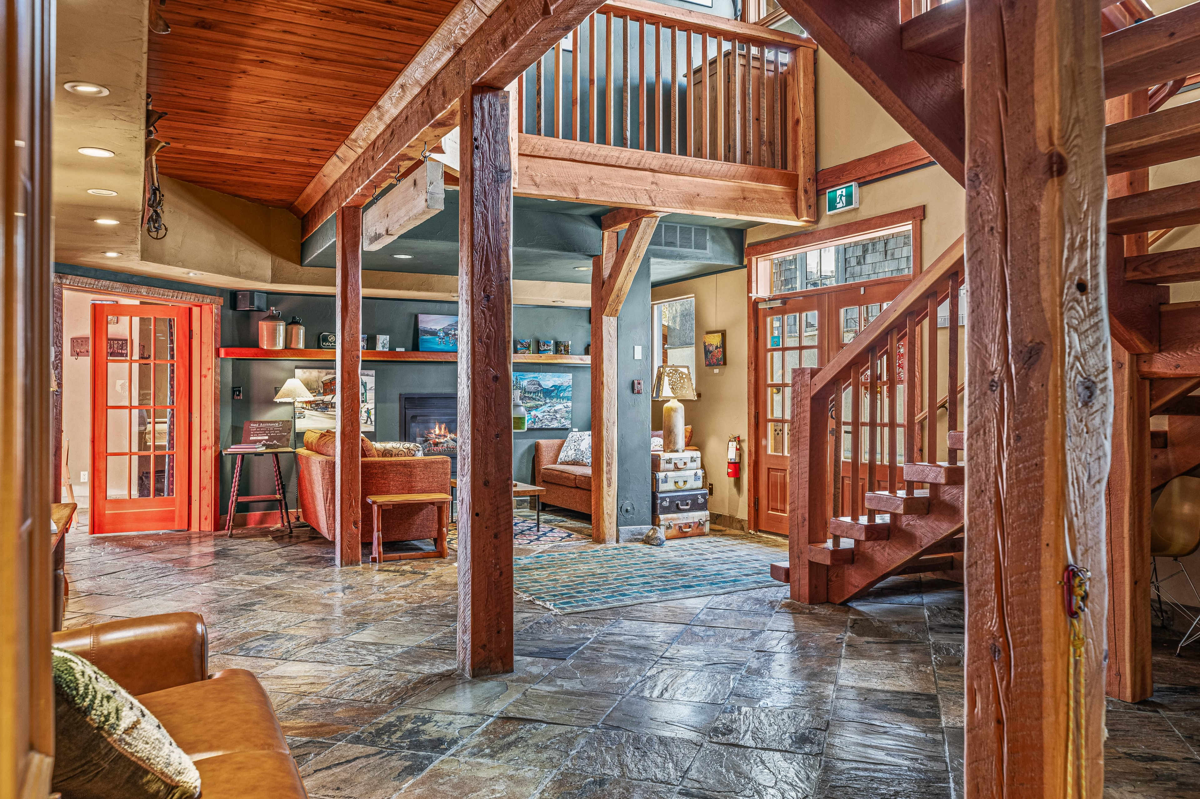 The image shows a spacious interior of a lodge-style living area with wooden beams, stone flooring, and a staircase leading to an upper level.