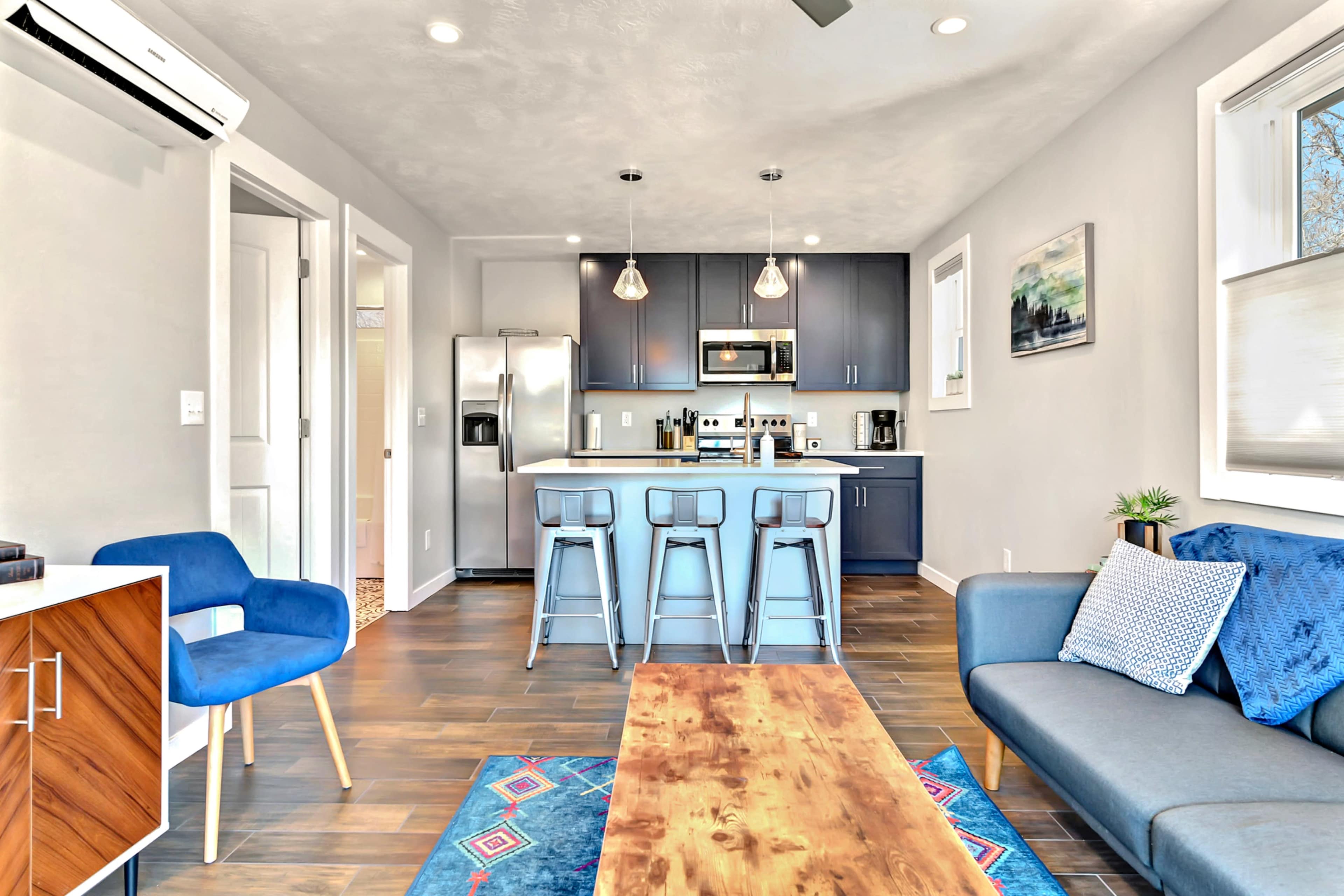 A modern living space featuring a kitchen with blue cabinetry, stainless steel appliances, and a wooden table surrounded by metal bar stools.