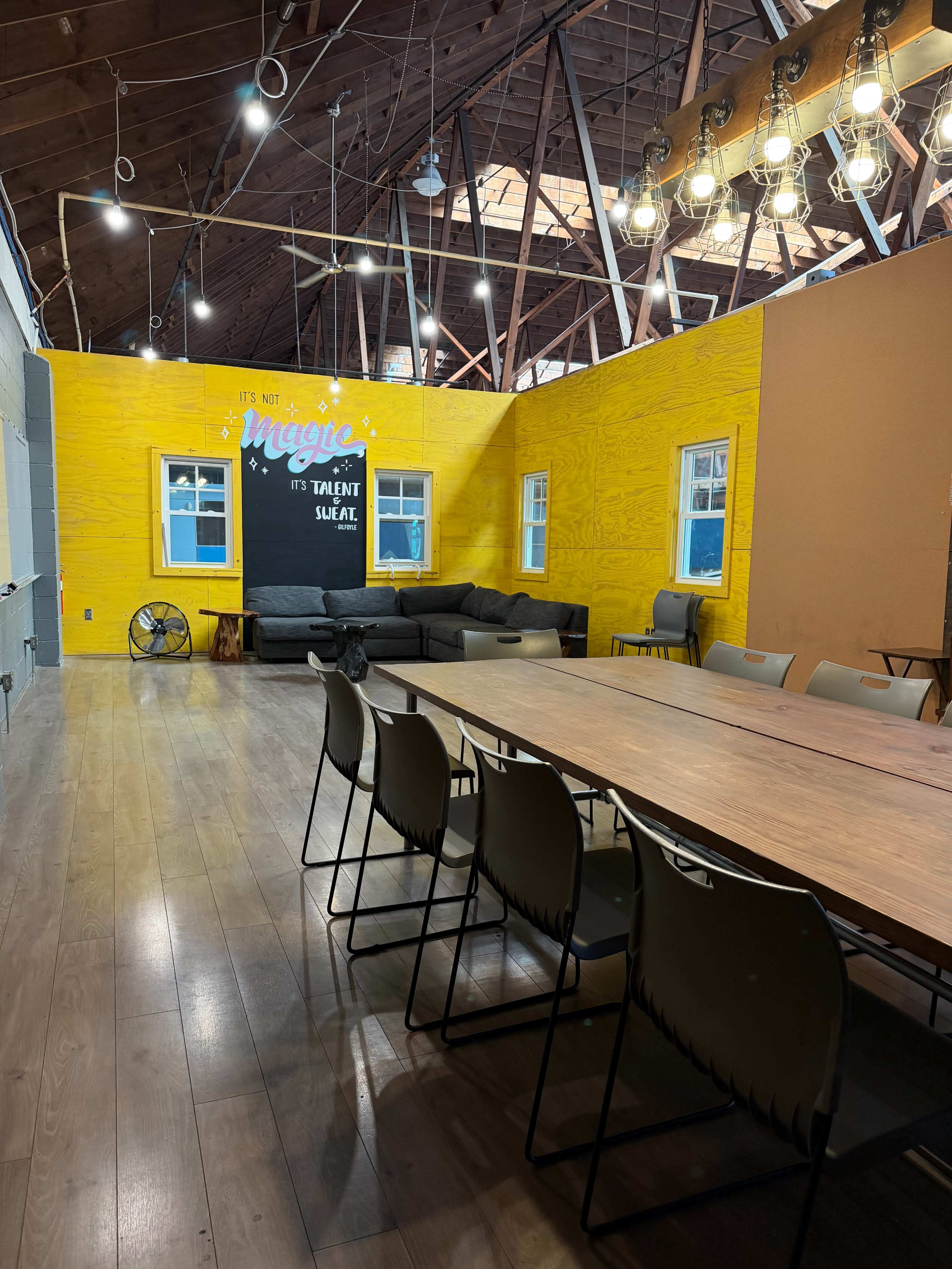 The image shows a bright yellow room with a long wooden table, six chairs, a sofa against the wall, and overhead lights in a rustic setting.