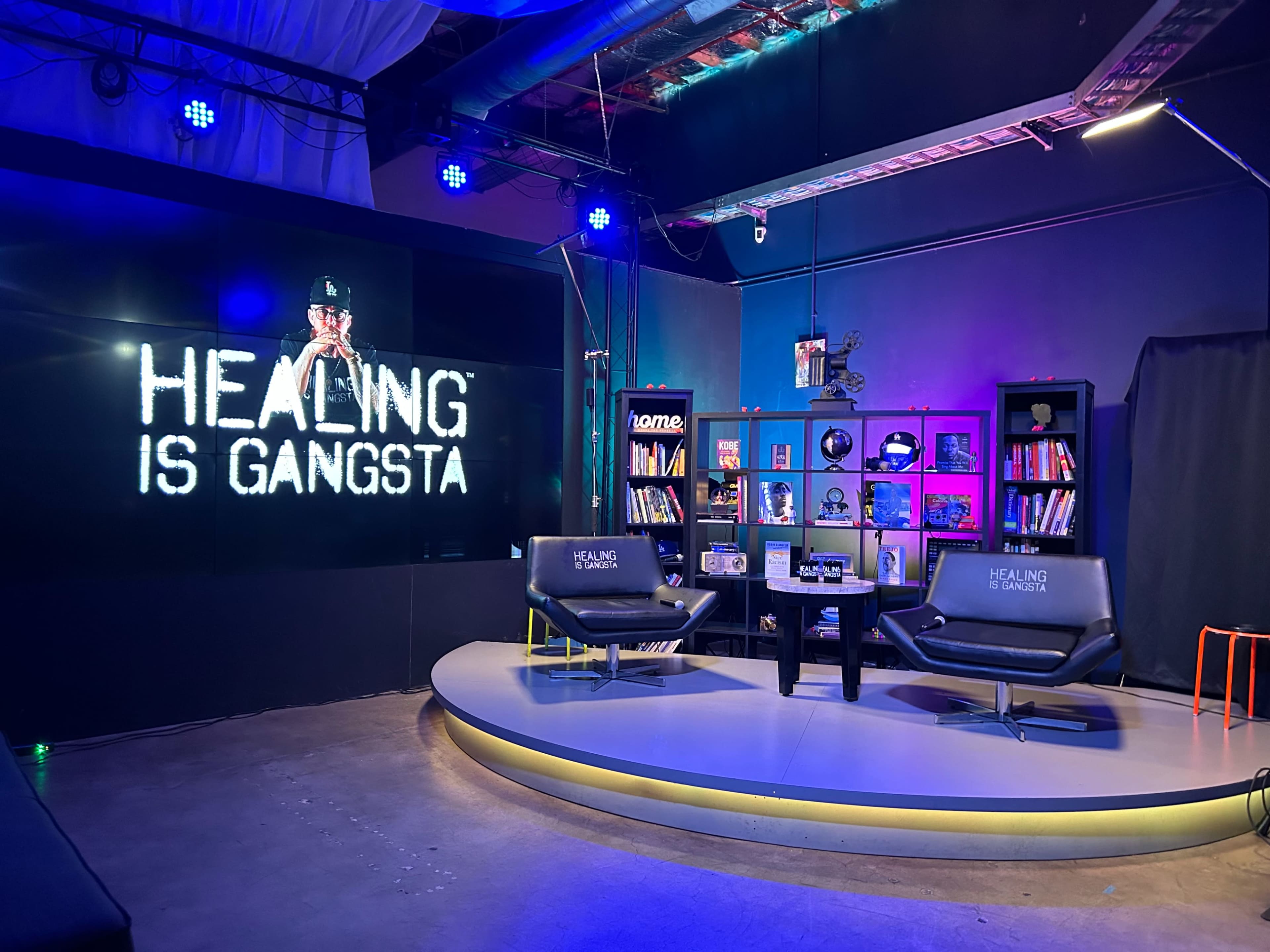 The image shows a modern studio set with two black chairs, a coffee table, and a large screen displaying the phrase "HEALING IS GANGSTA."