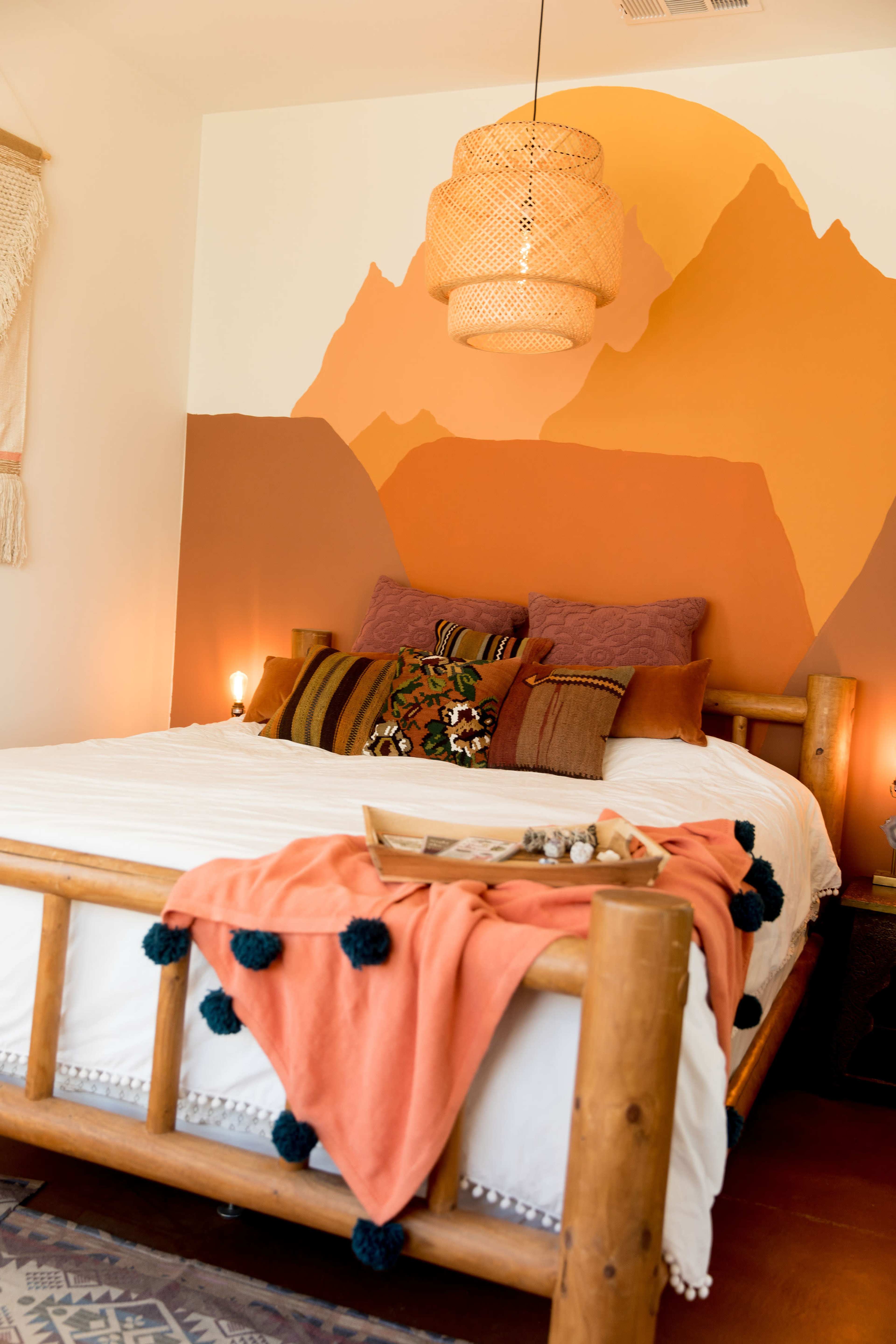 A wooden bed adorned with colorful pillows and a textured blanket is set against a mountain-themed mural in warm tones.