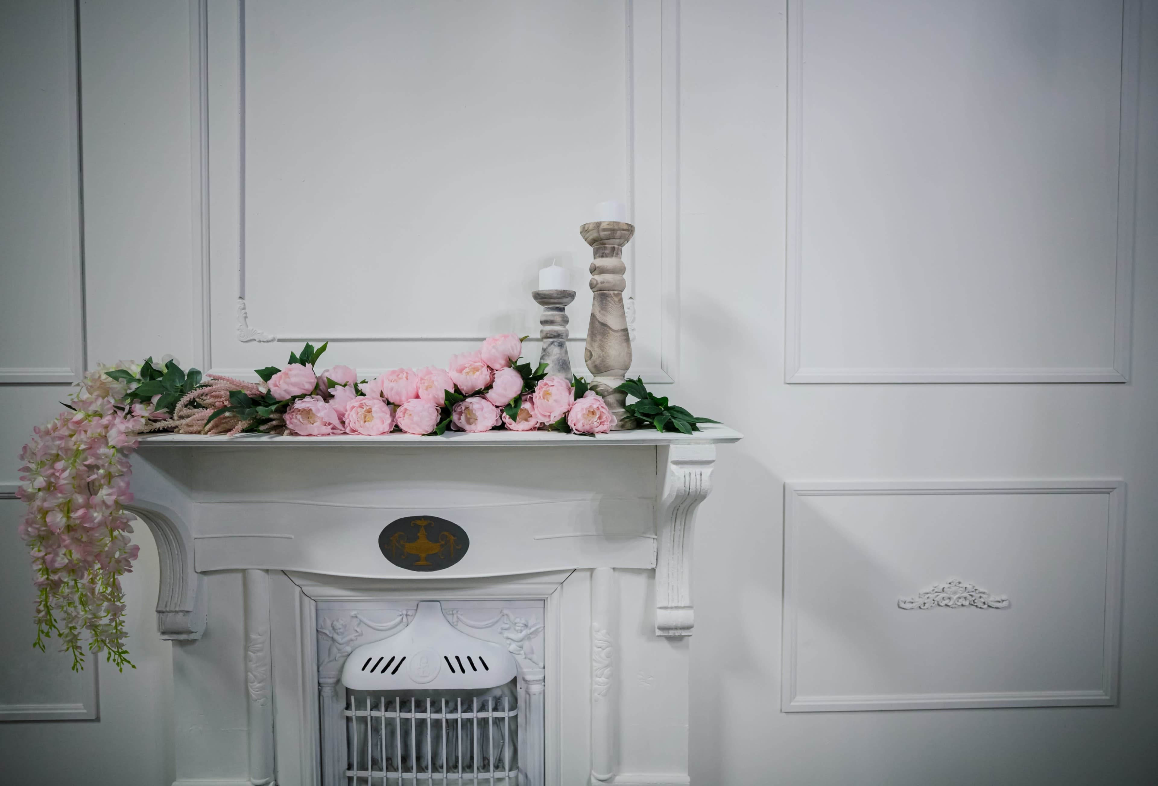 A white mantel adorned with pink flowers and two decorative candles against a plain wall.