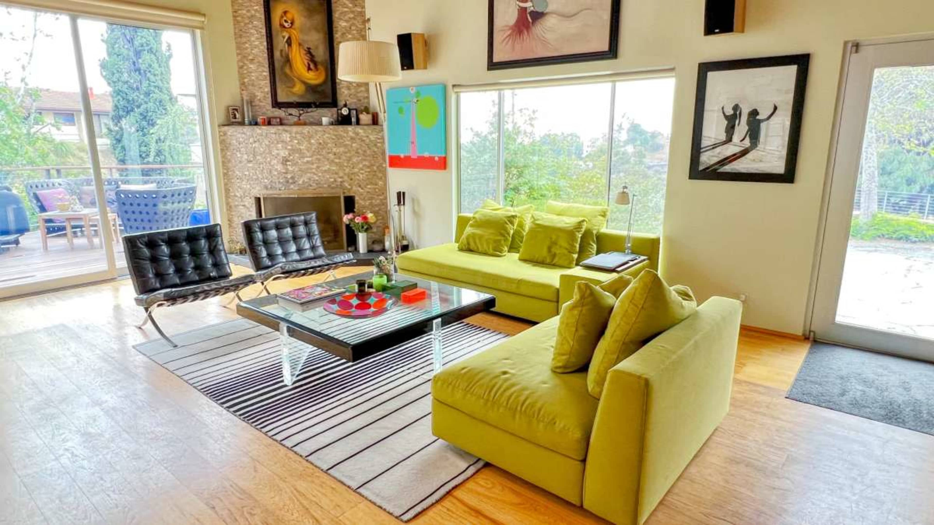 The image shows a modern living room with a yellow sectional sofa, a glass coffee table, and large windows overlooking a garden.