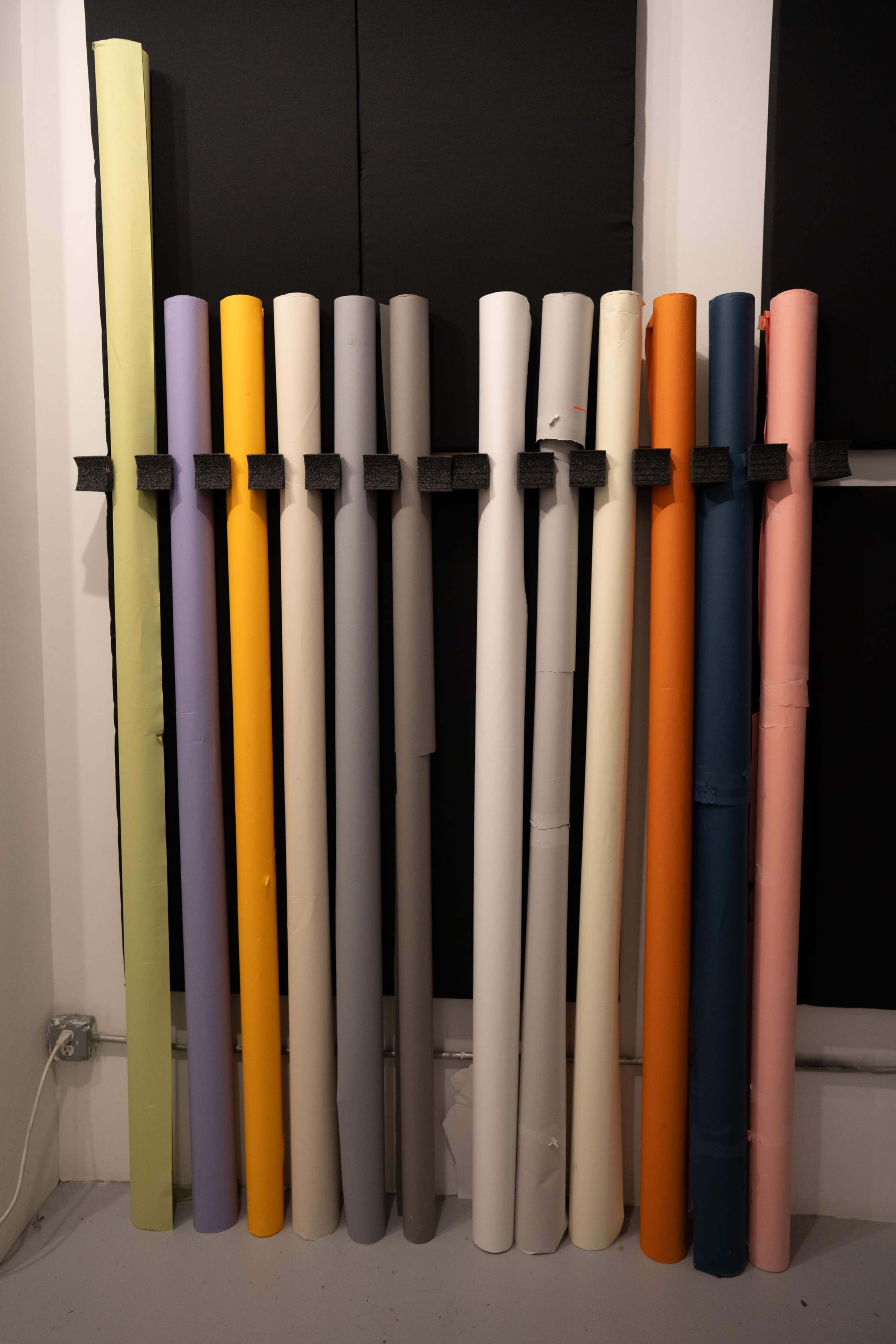 The image shows a collection of rolled paper in various colors, arranged vertically against a wall.