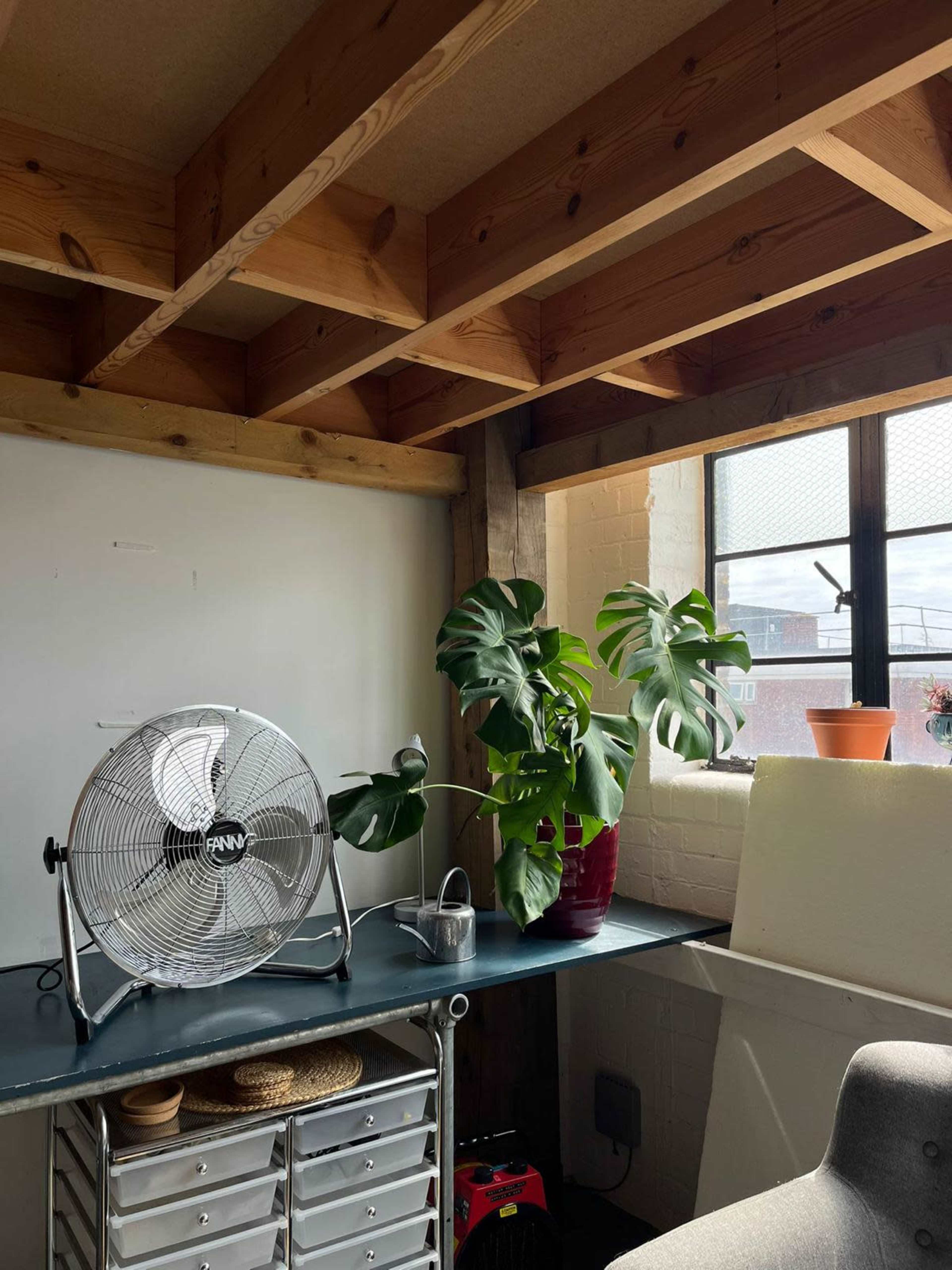 The image shows a workspace with a metal fan, a potted monstera plant, and shelves against a wall with exposed wooden beams overhead.