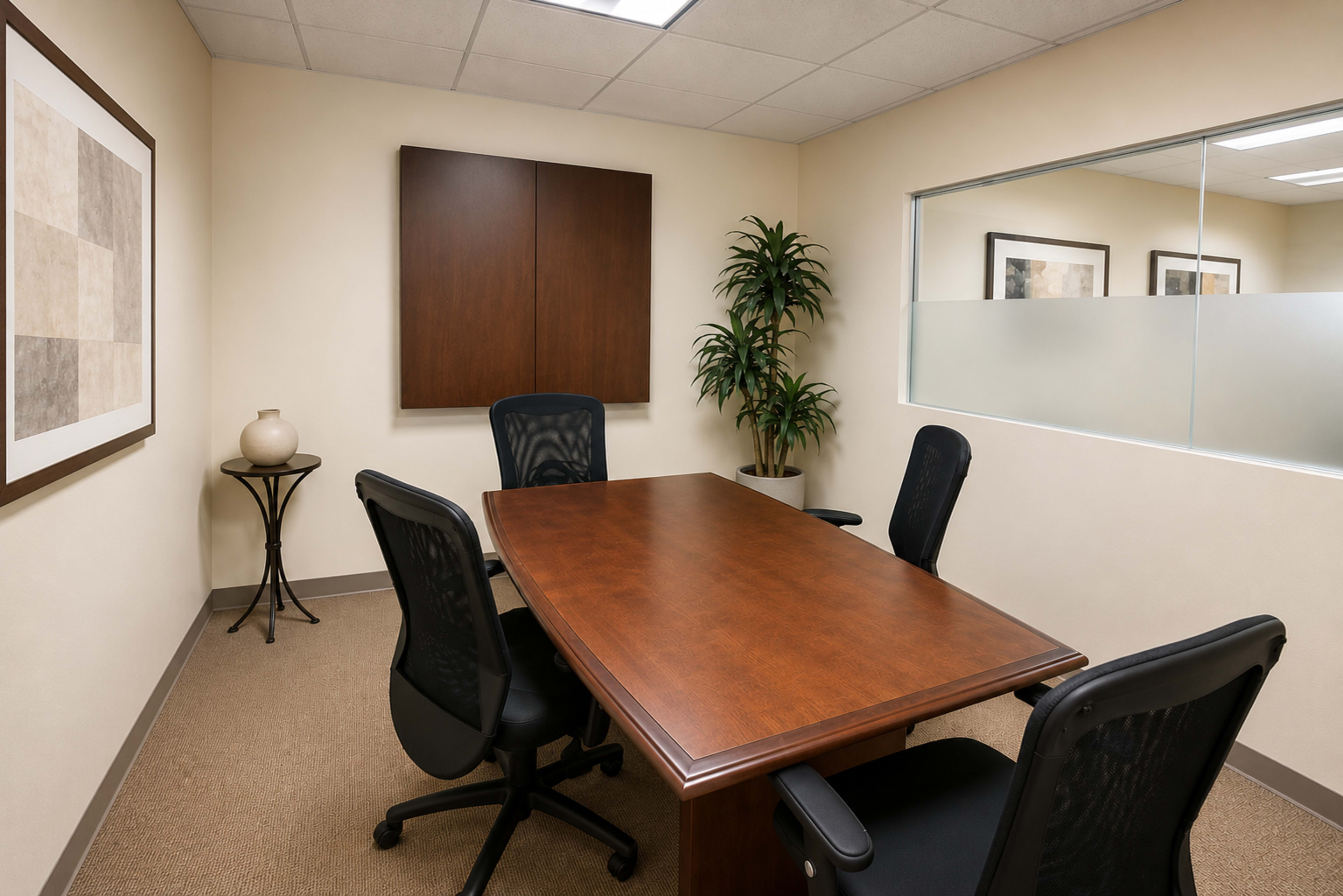 The image shows a small conference room featuring a brown wooden table, four black office chairs, a plant in the corner, and framed artwork on the walls.