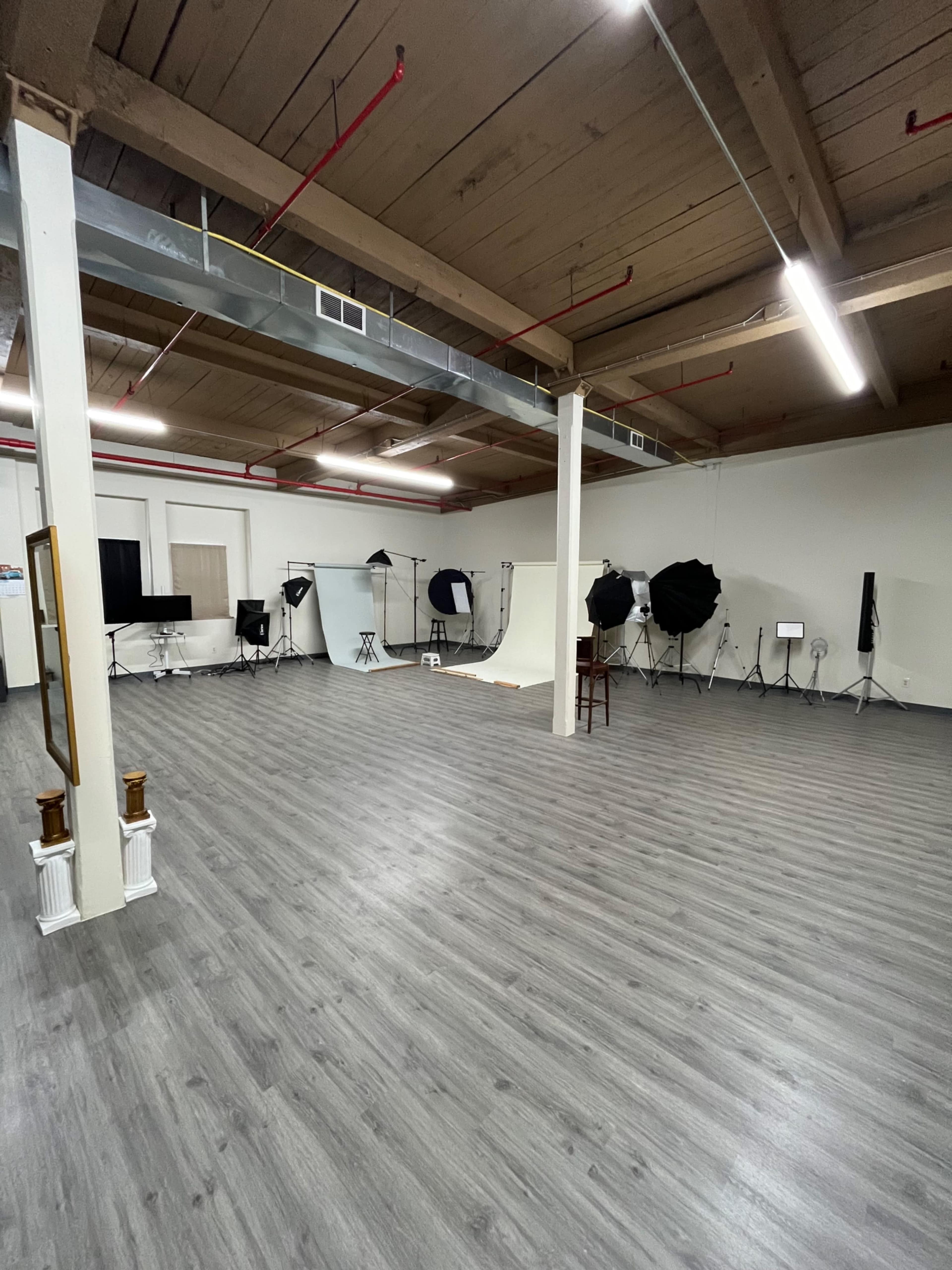 The image shows a spacious photography studio with multiple lighting setups and background equipment arranged around a smooth, wooden floor.