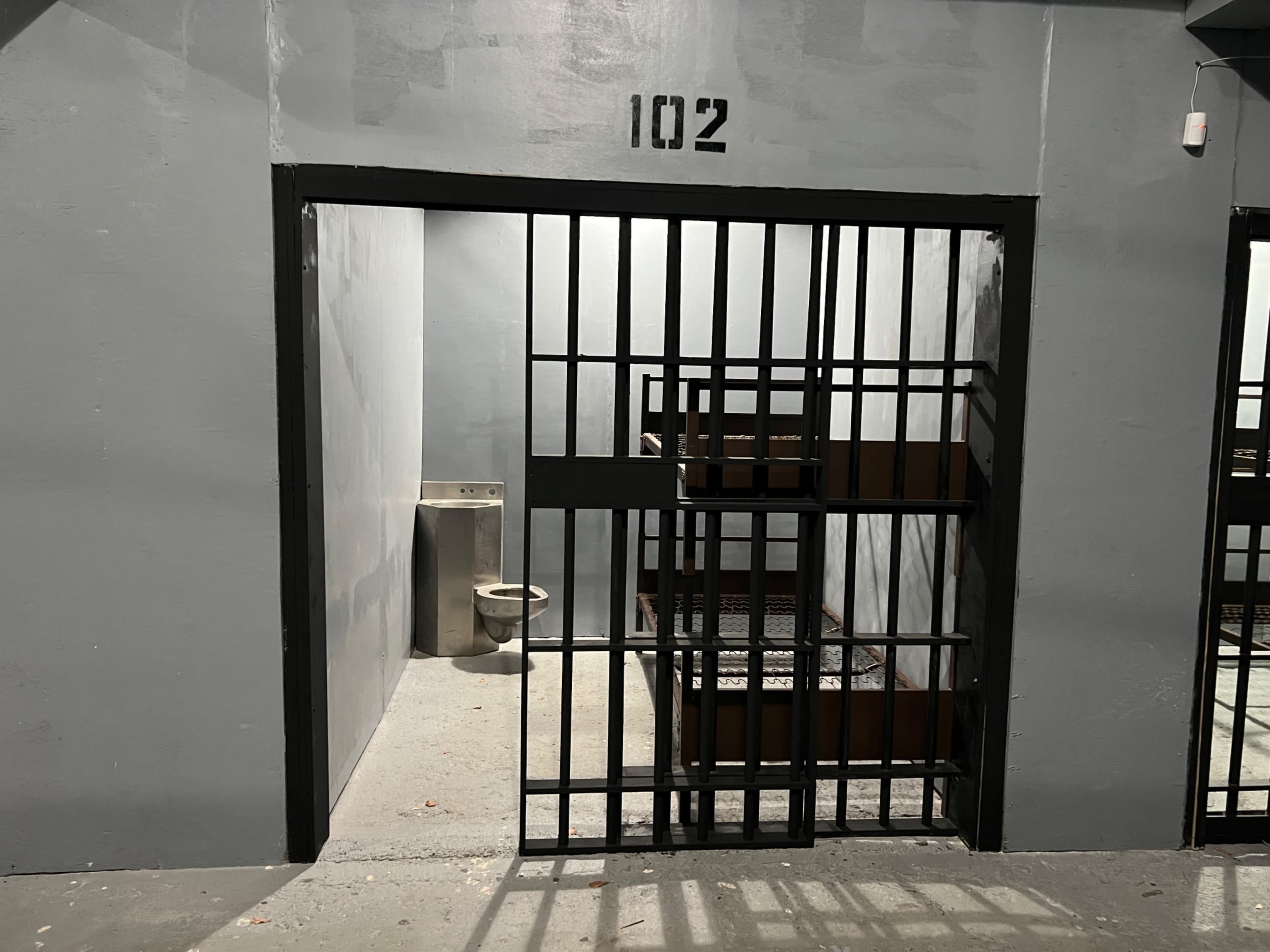An open jail cell marked with the number 102, featuring a toilet and two metal bunks.