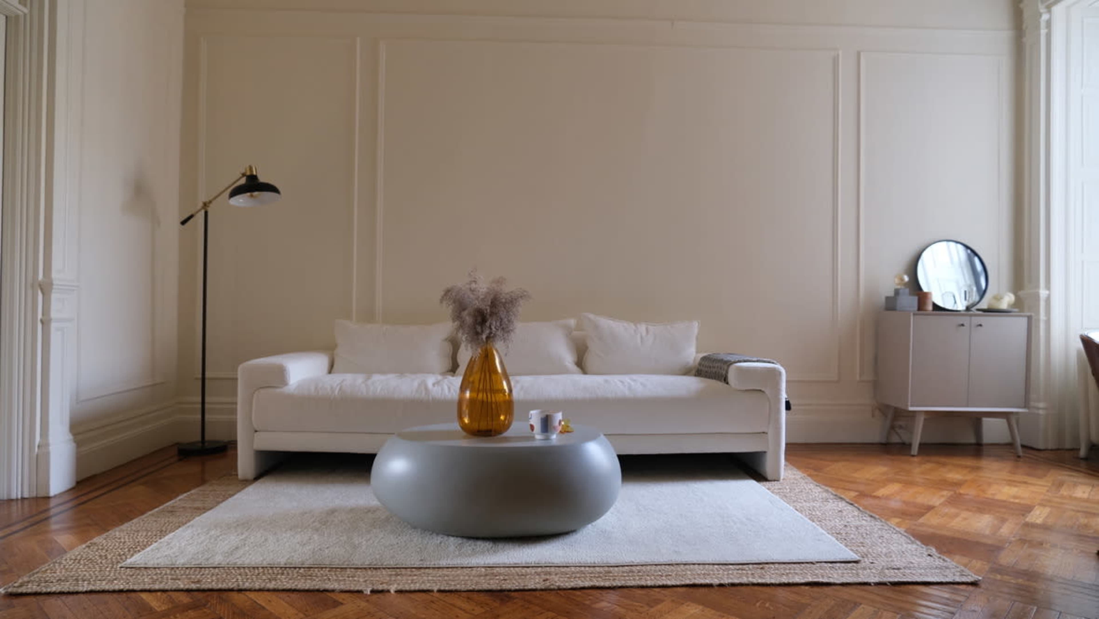 A modern living room features a white sofa, a round gray coffee table with a yellow vase, and wooden flooring.
