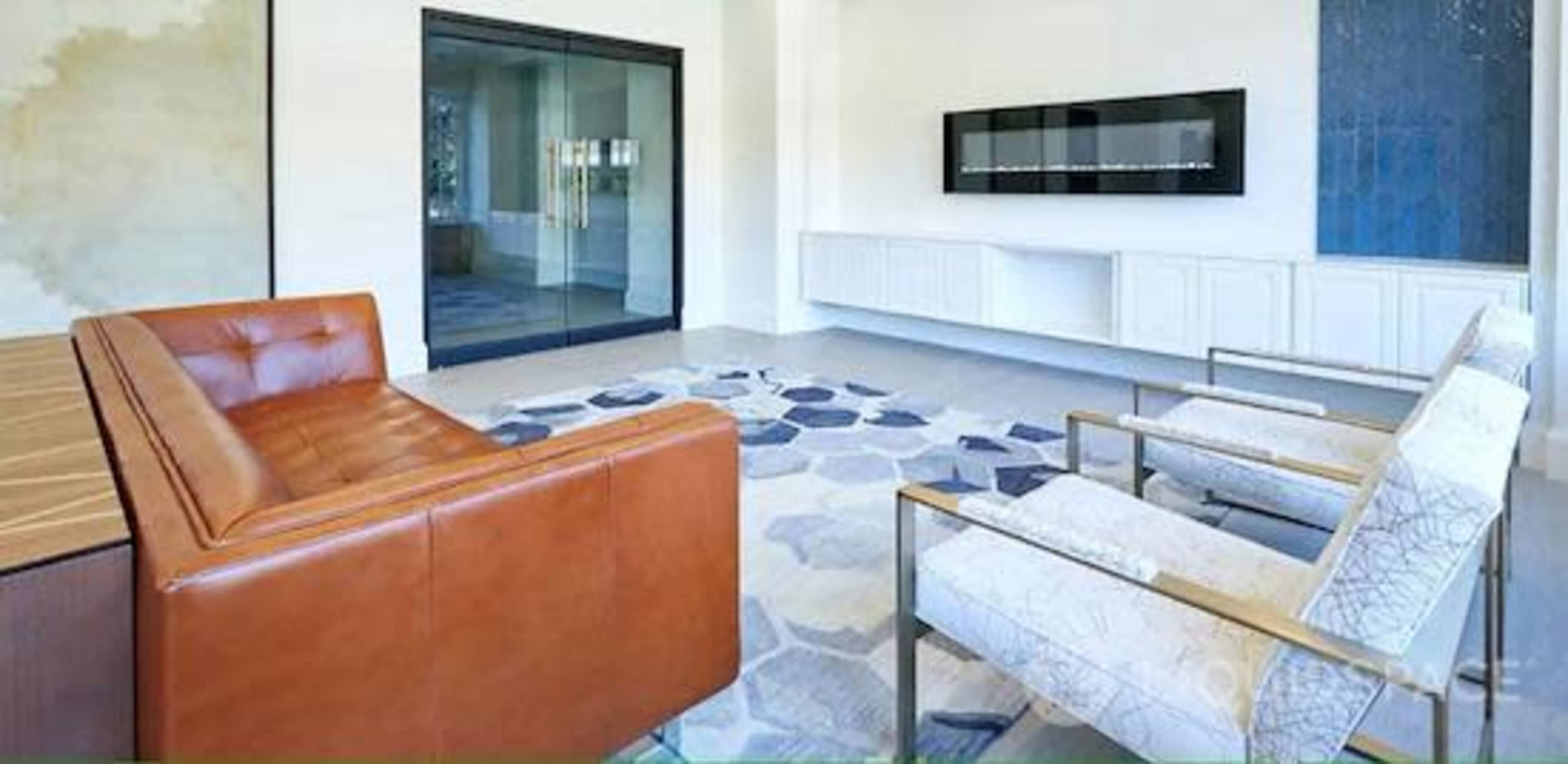 A modern, minimalist interior with a brown leather sofa, two white armchairs, and a patterned rug in a well-lit room featuring a glass door and a wall-mounted television.
