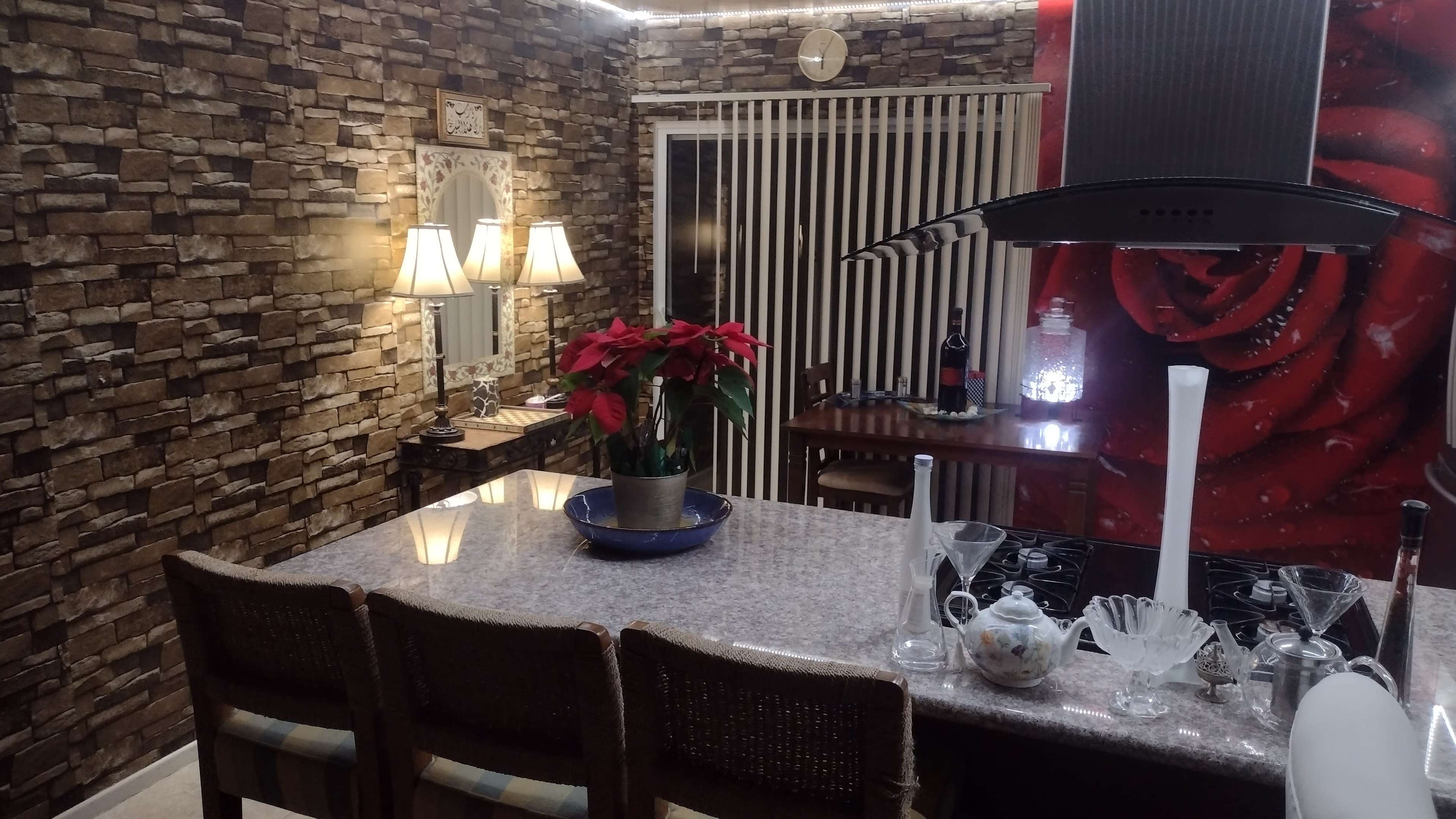 A kitchen with stone-patterned walls, a granite countertop with three stools, and a decorative arrangement of flowers and glassware.
