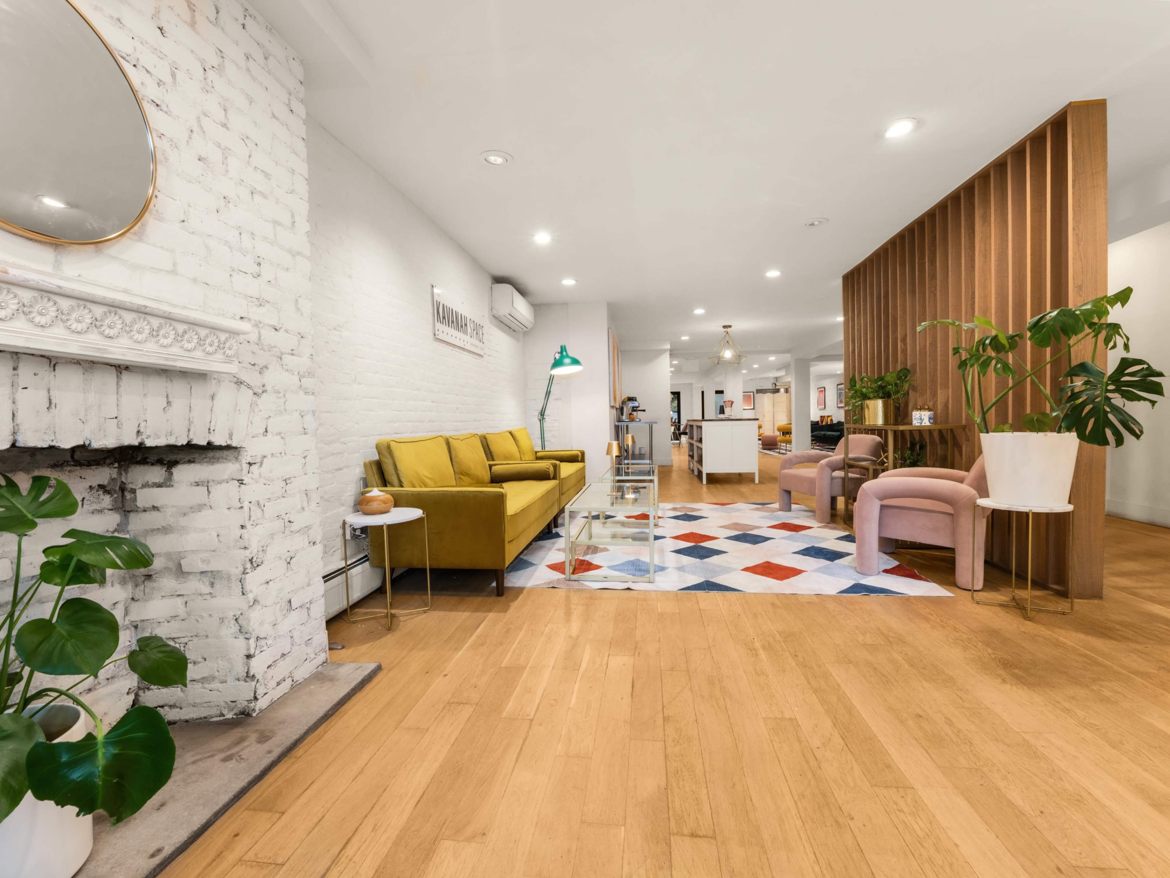The image shows a modern interior living space featuring a yellow sofa, pink chairs, a geometric rug, and a brick accent wall.