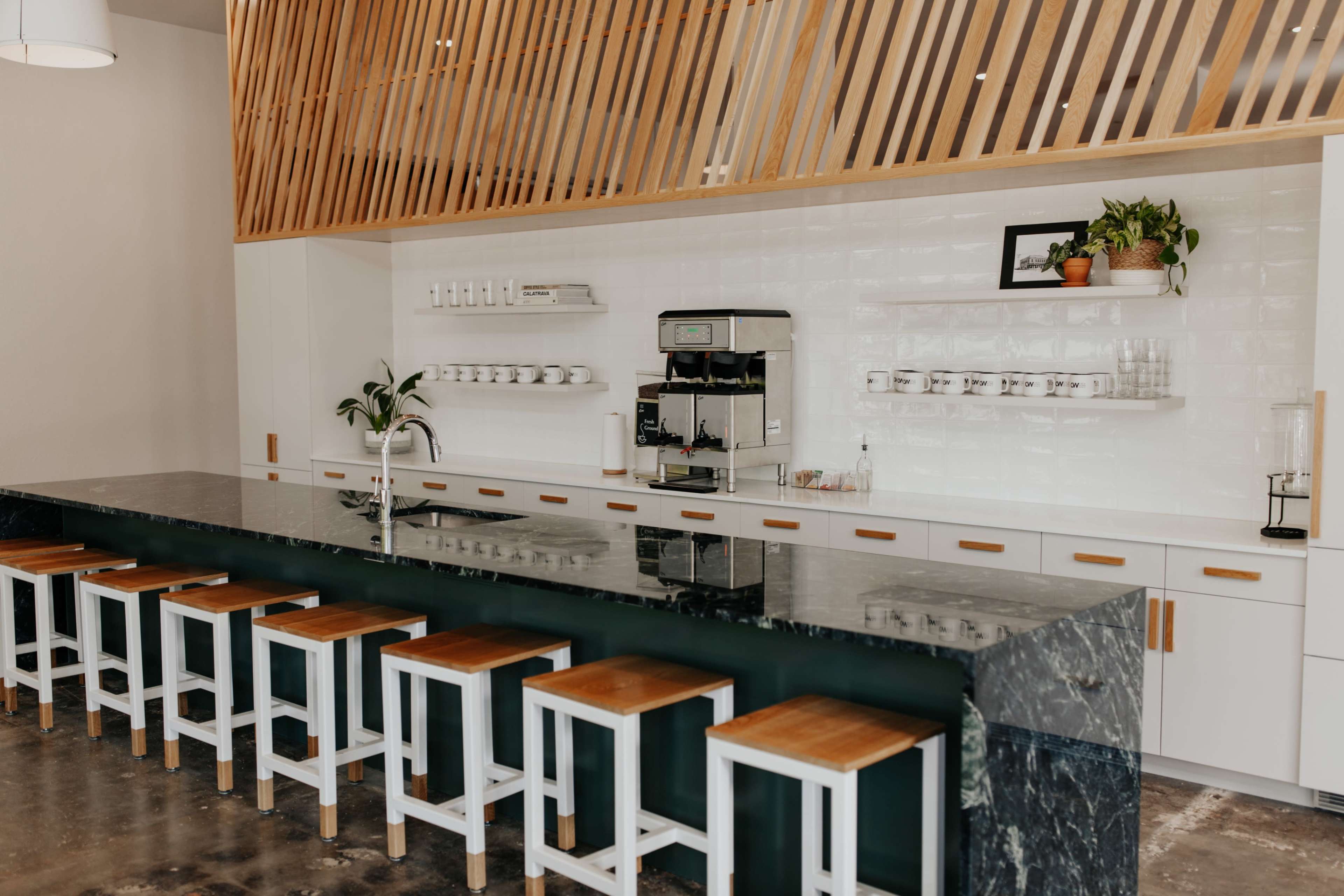 A modern cafe interior features a long marble counter with wooden stools, a coffee machine, and neatly arranged shelves above.