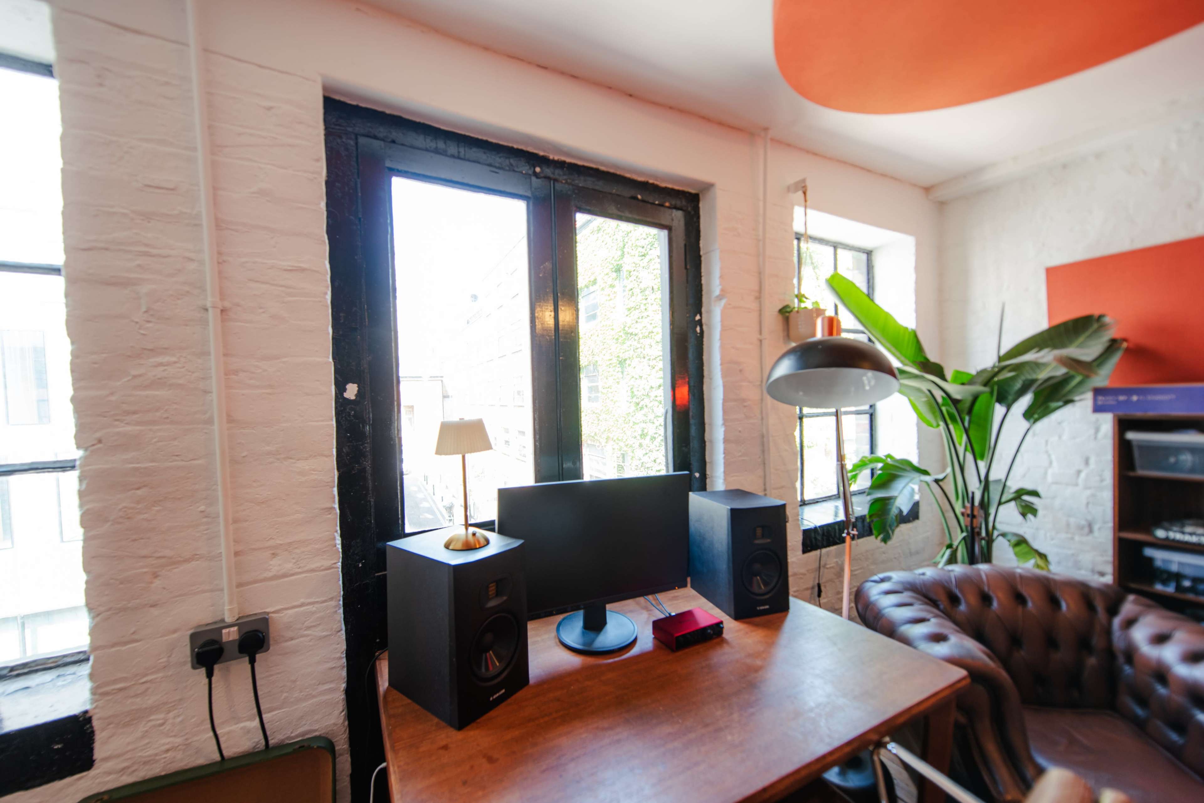 The image shows a cozy room with a desk featuring two speakers and a monitor, large windows allowing natural light, and a plant in the corner.