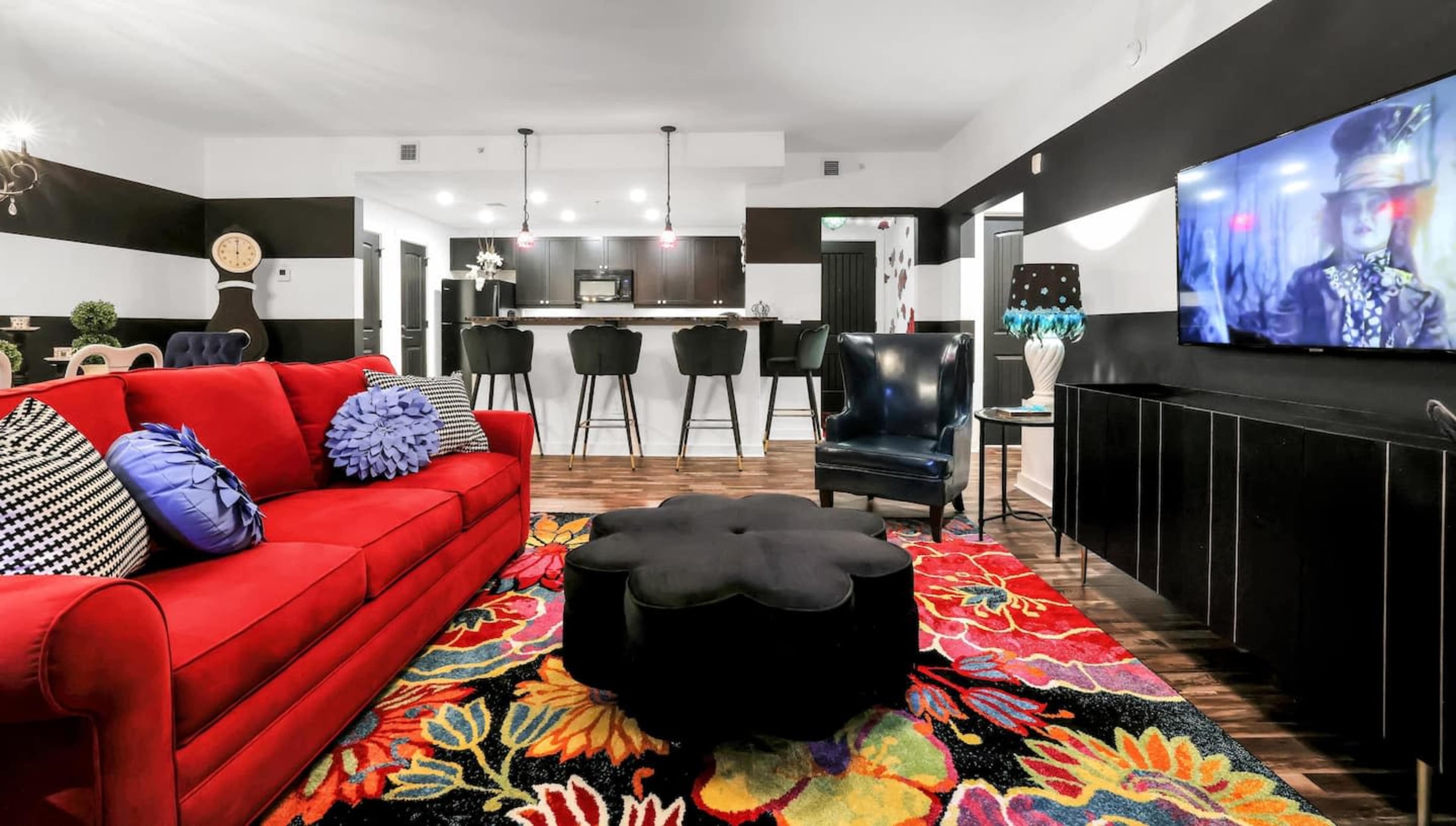 A colorful living room featuring a red sofa, a black flower-shaped coffee table, and a large television on one wall, with a kitchen area visible in the background.