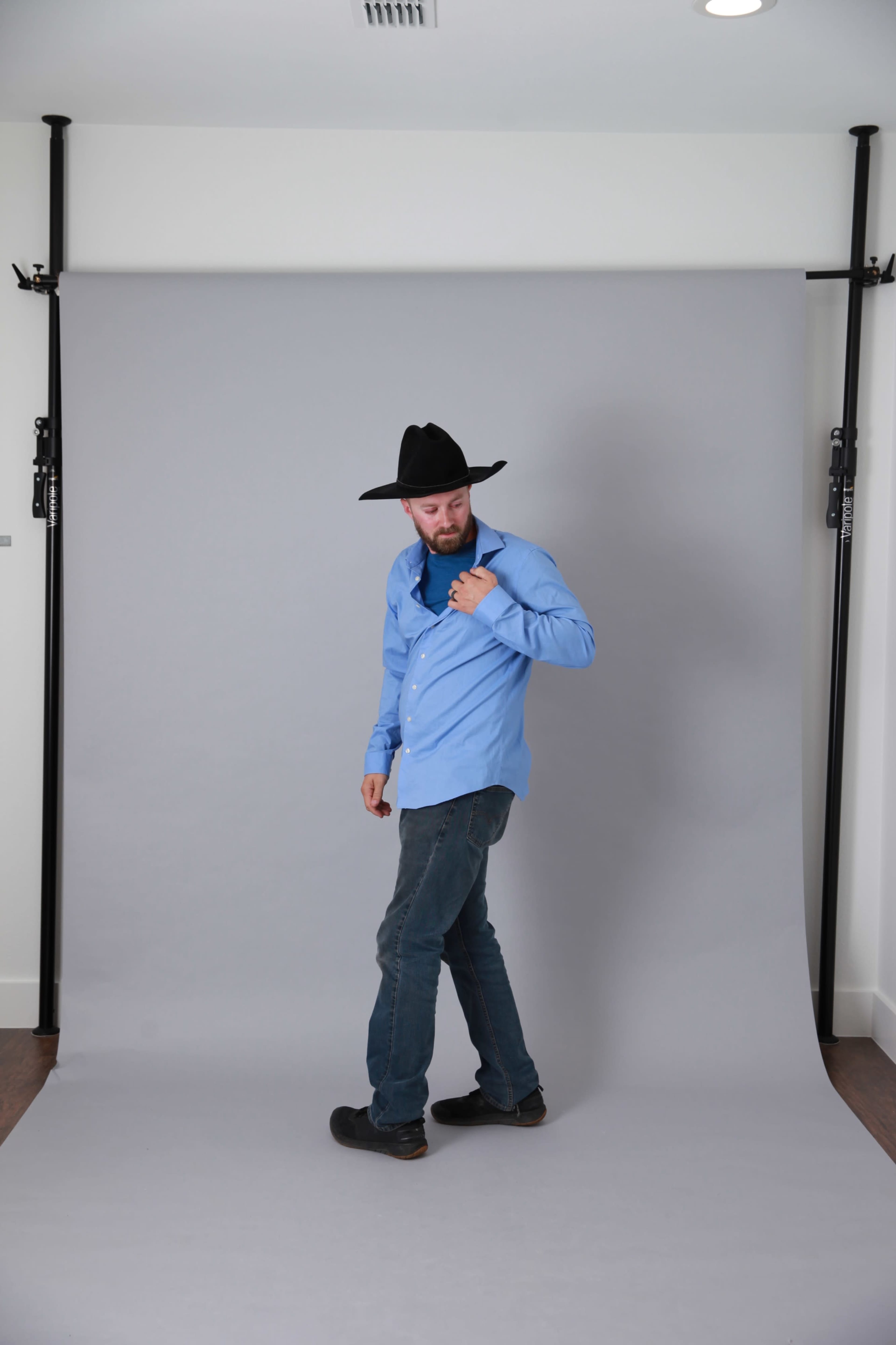 A man wearing a blue shirt and black hat stands in front of a gray backdrop, adjusting his shirt with one hand.