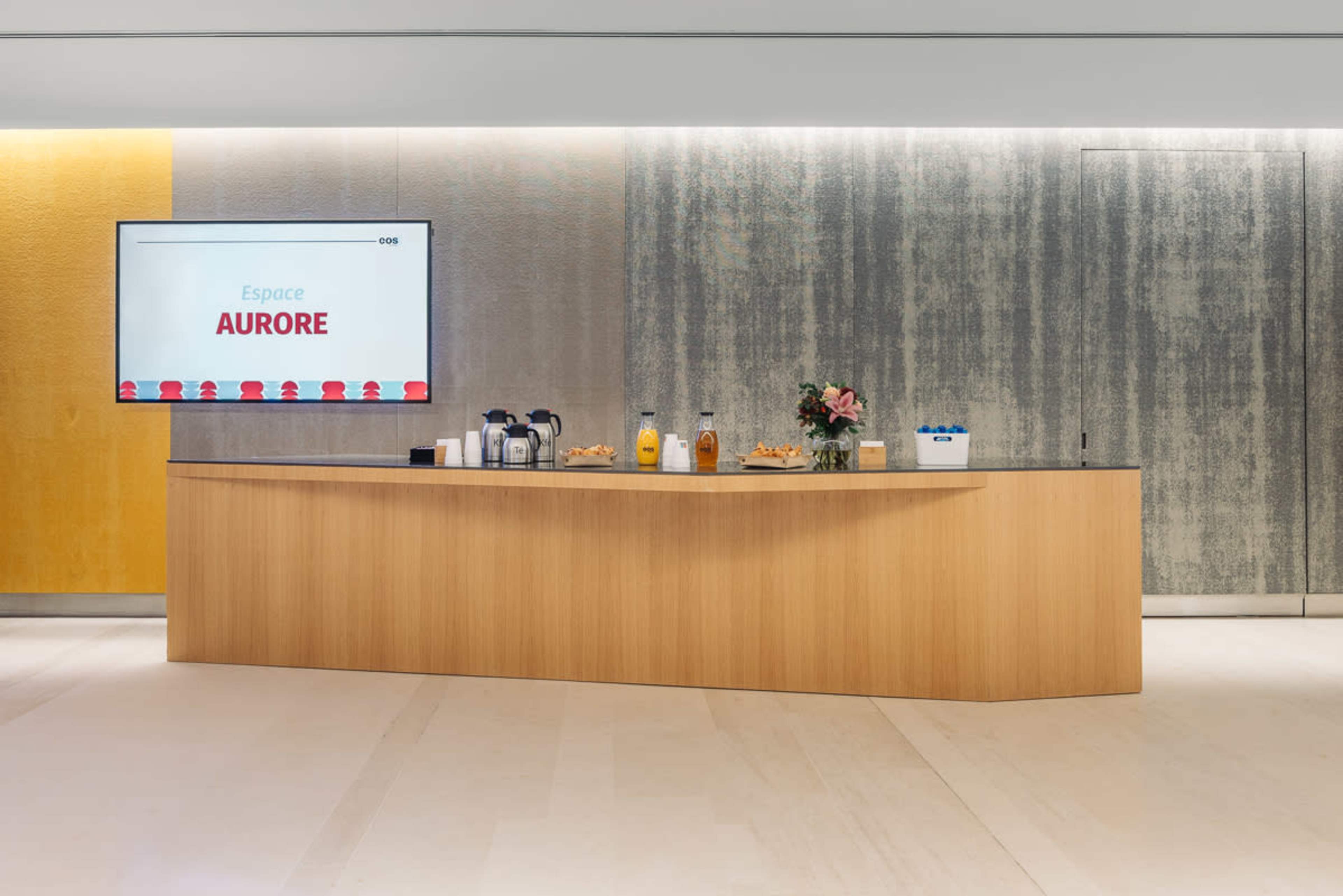 A wooden reception desk with refreshments and a display screen showing "Espace Aurore" is set against a textured wall.
