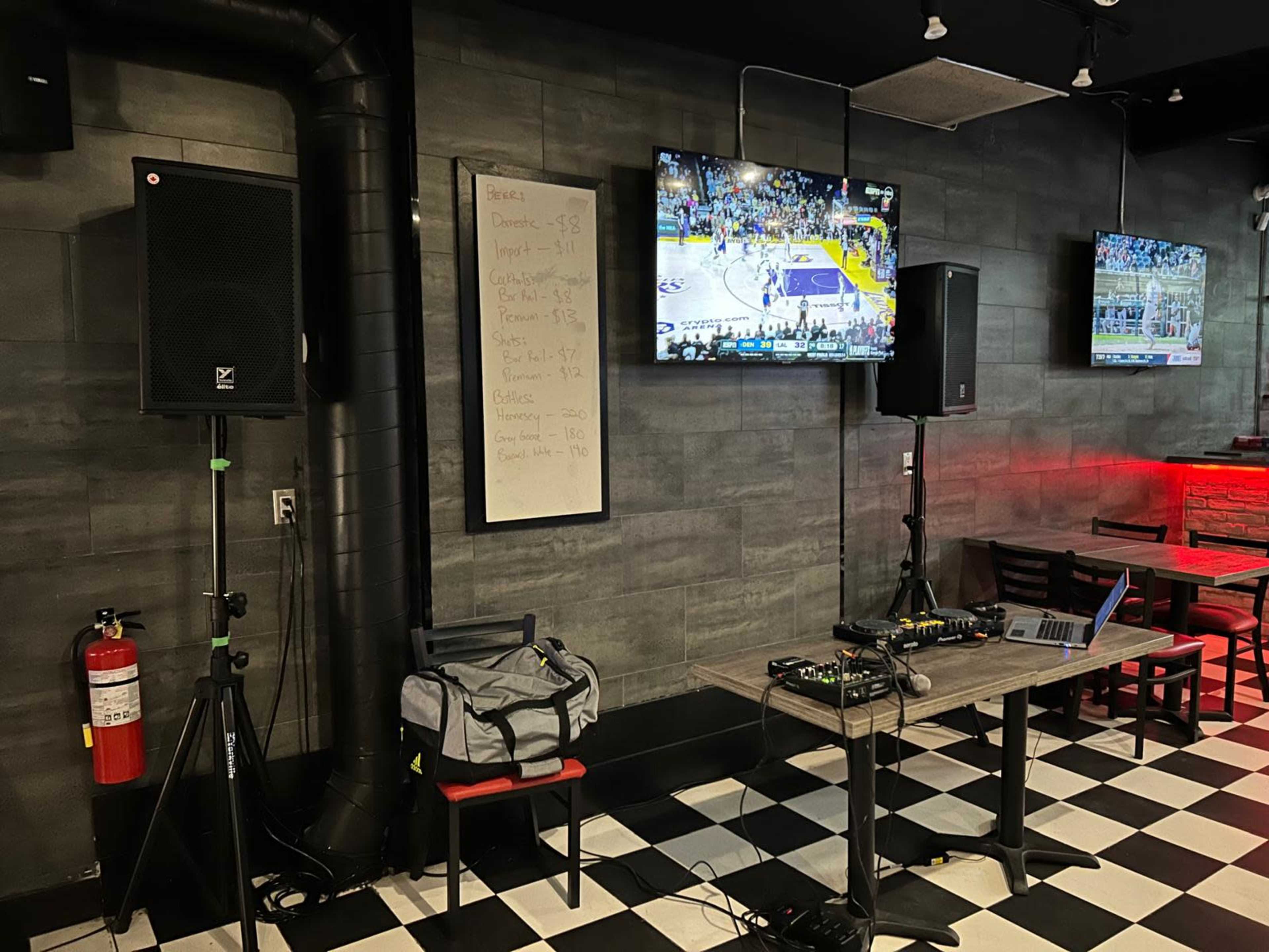 Two televisions display a basketball game, while audio equipment and a laptop are set up on a table in a venue with a checkered floor.