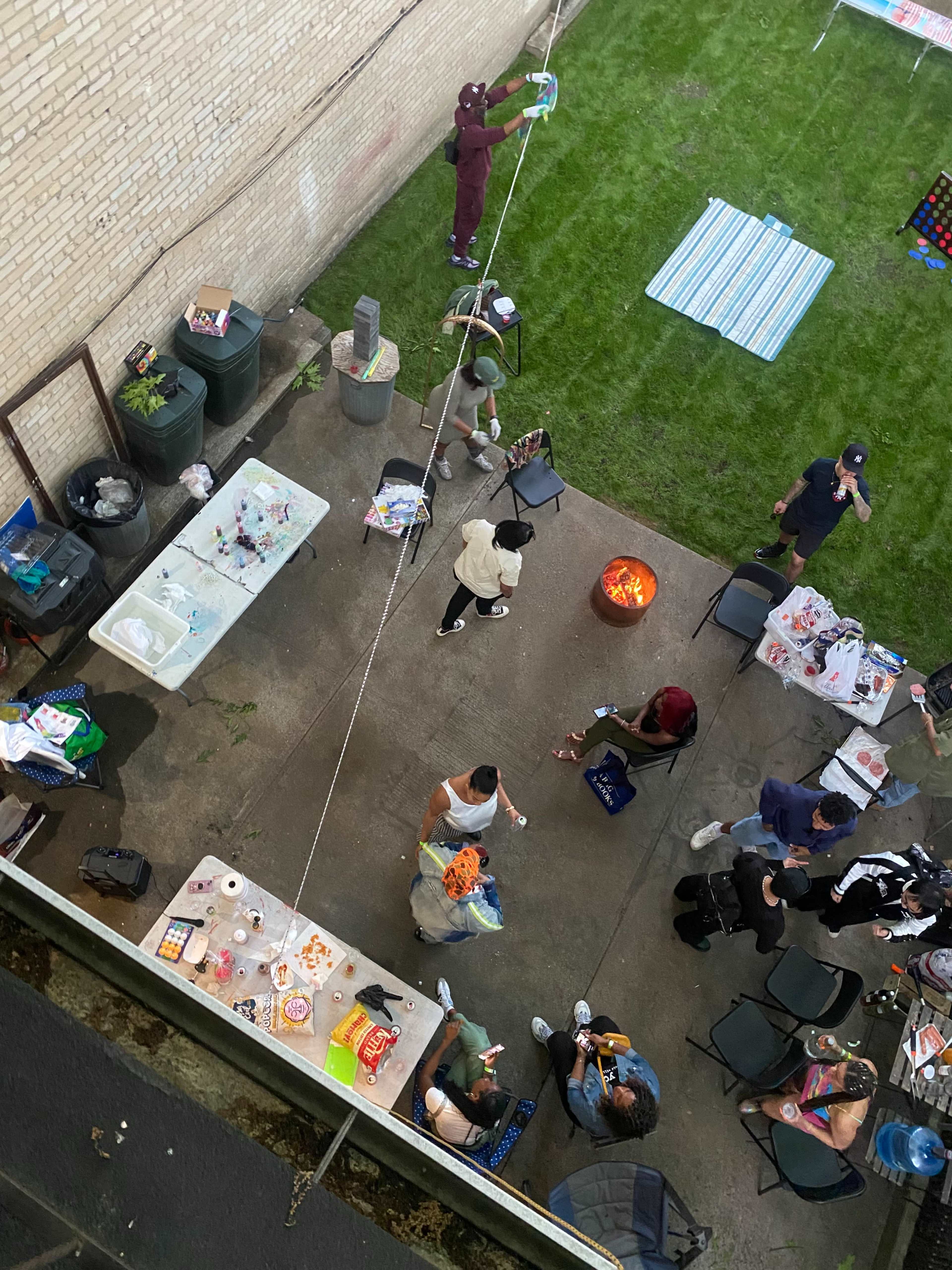 A group of people gathers in an outdoor space with a fire pit, tables filled with food, and various activities in progress.