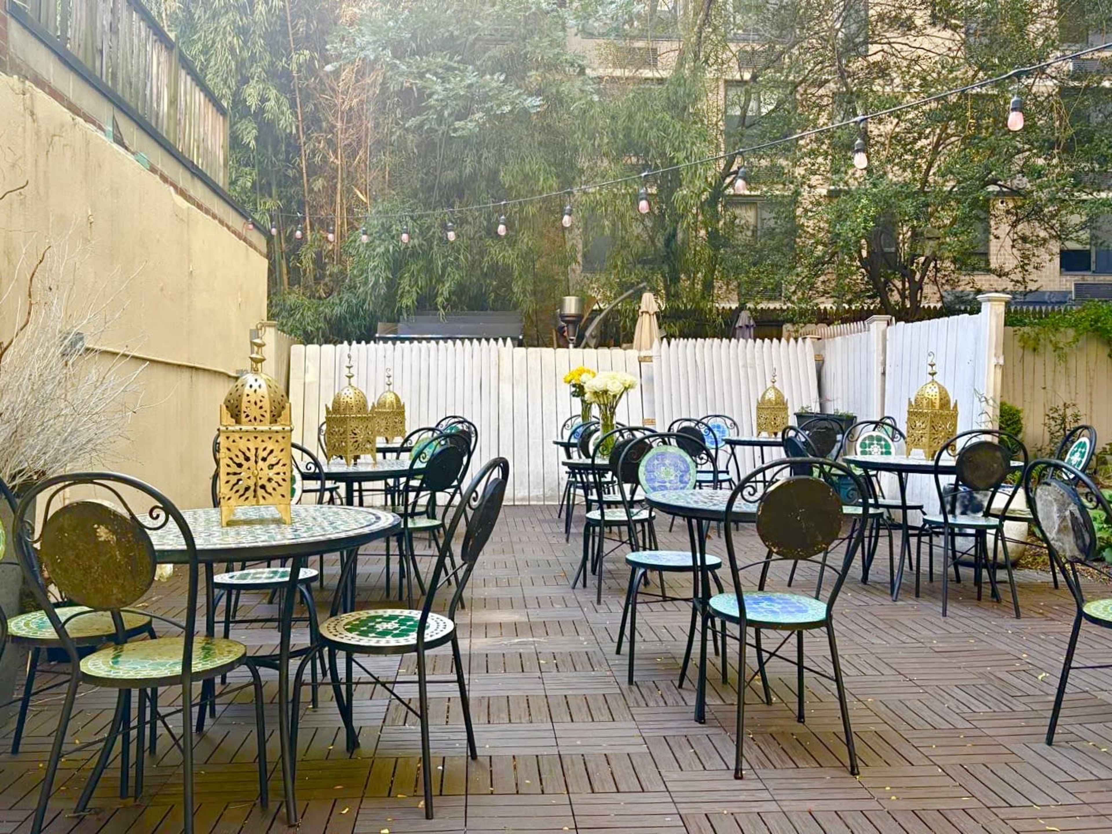 The image shows an outdoor patio area with several round tables and chairs arranged around a wooden deck, decorated with lanterns and surrounded by greenery.