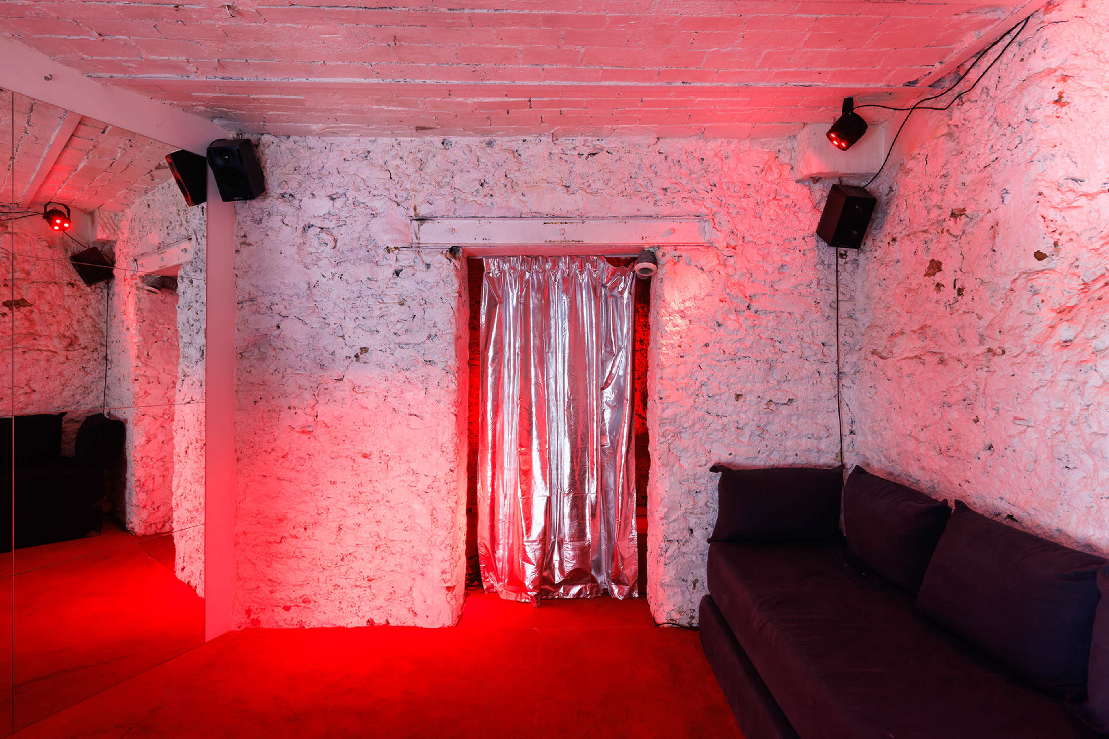 The image depicts a room with textured white walls, red lighting, a silver curtain, and a dark couch against one side.