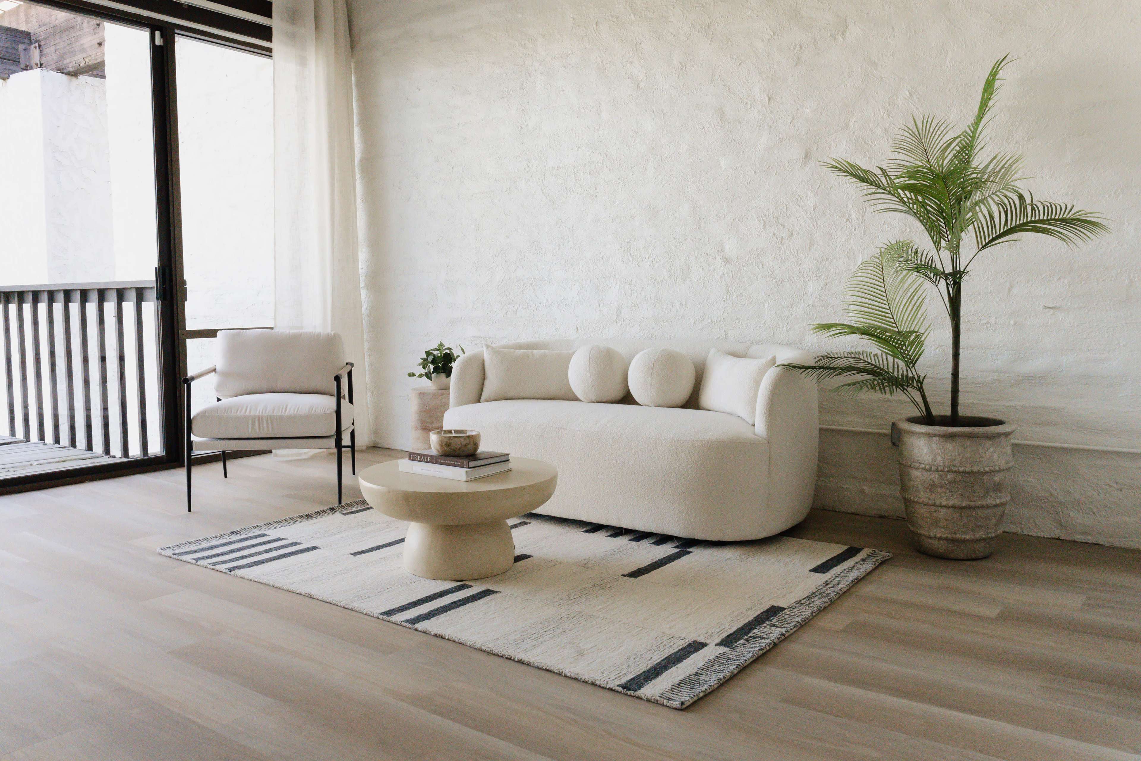 The image shows a minimalist living area featuring a soft, curved sofa, a round coffee table, a chair with a metal frame, a potted plant, and a patterned rug on a light wooden floor.