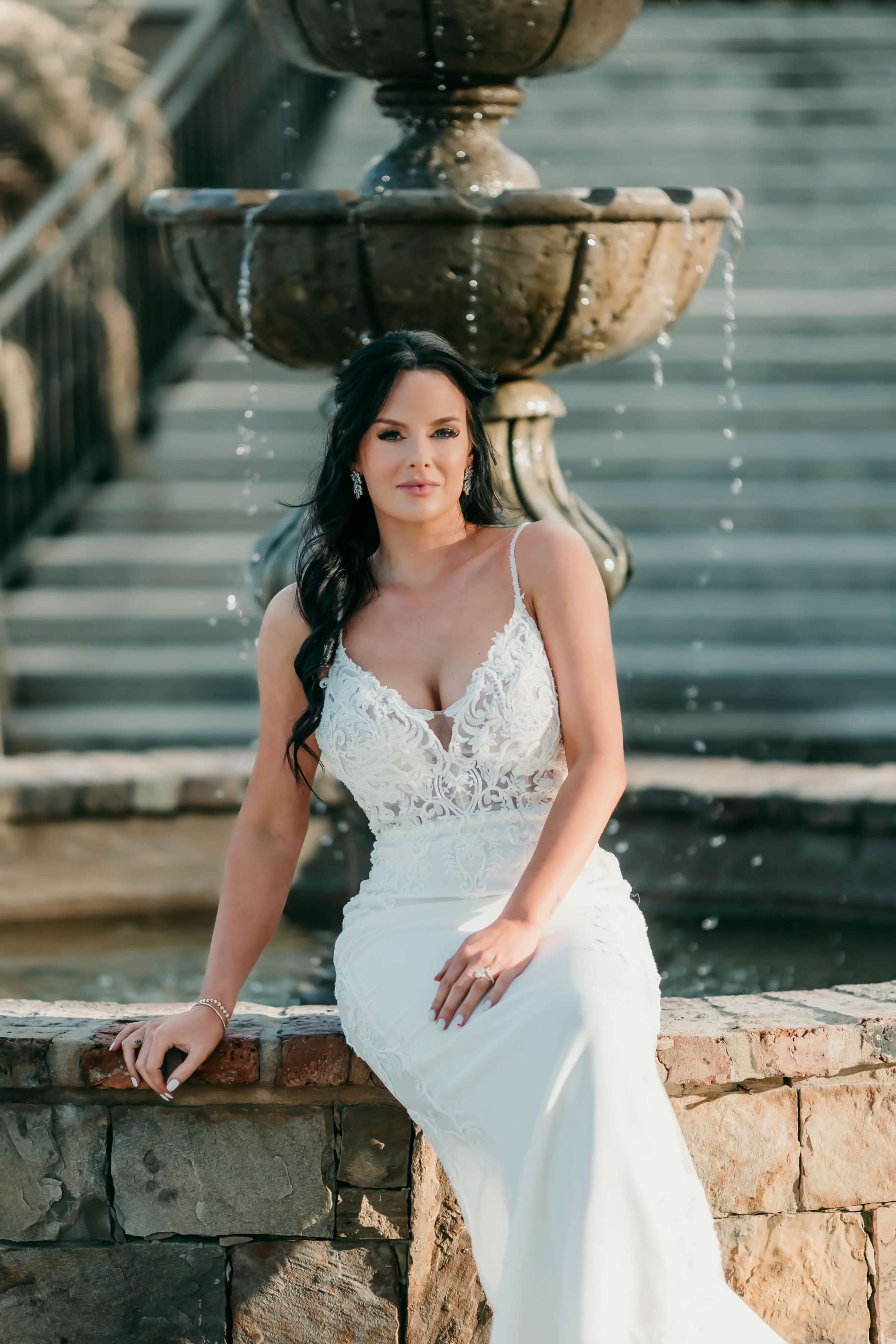 A woman in a white dress sits by a stone fountain with water flowing gently around her.