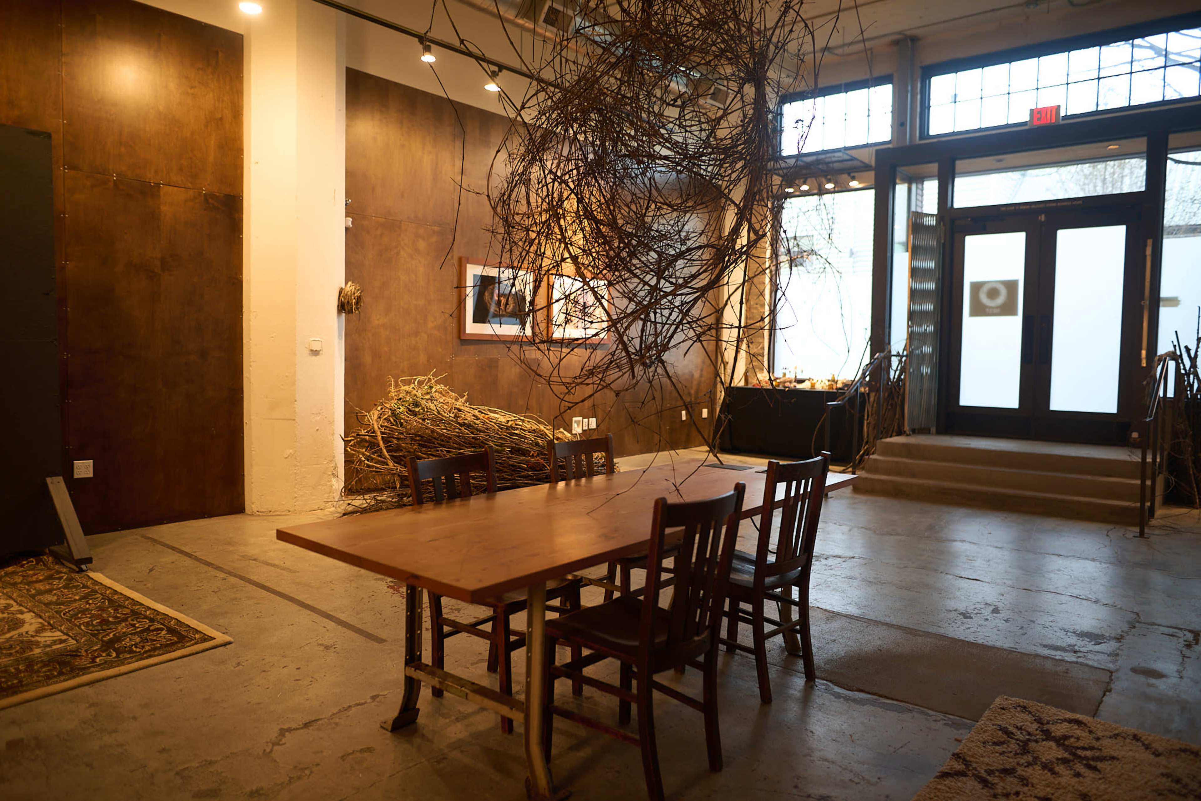The image shows an interior space with a wooden table surrounded by chairs, an art display on the walls, and dried branches hanging above.